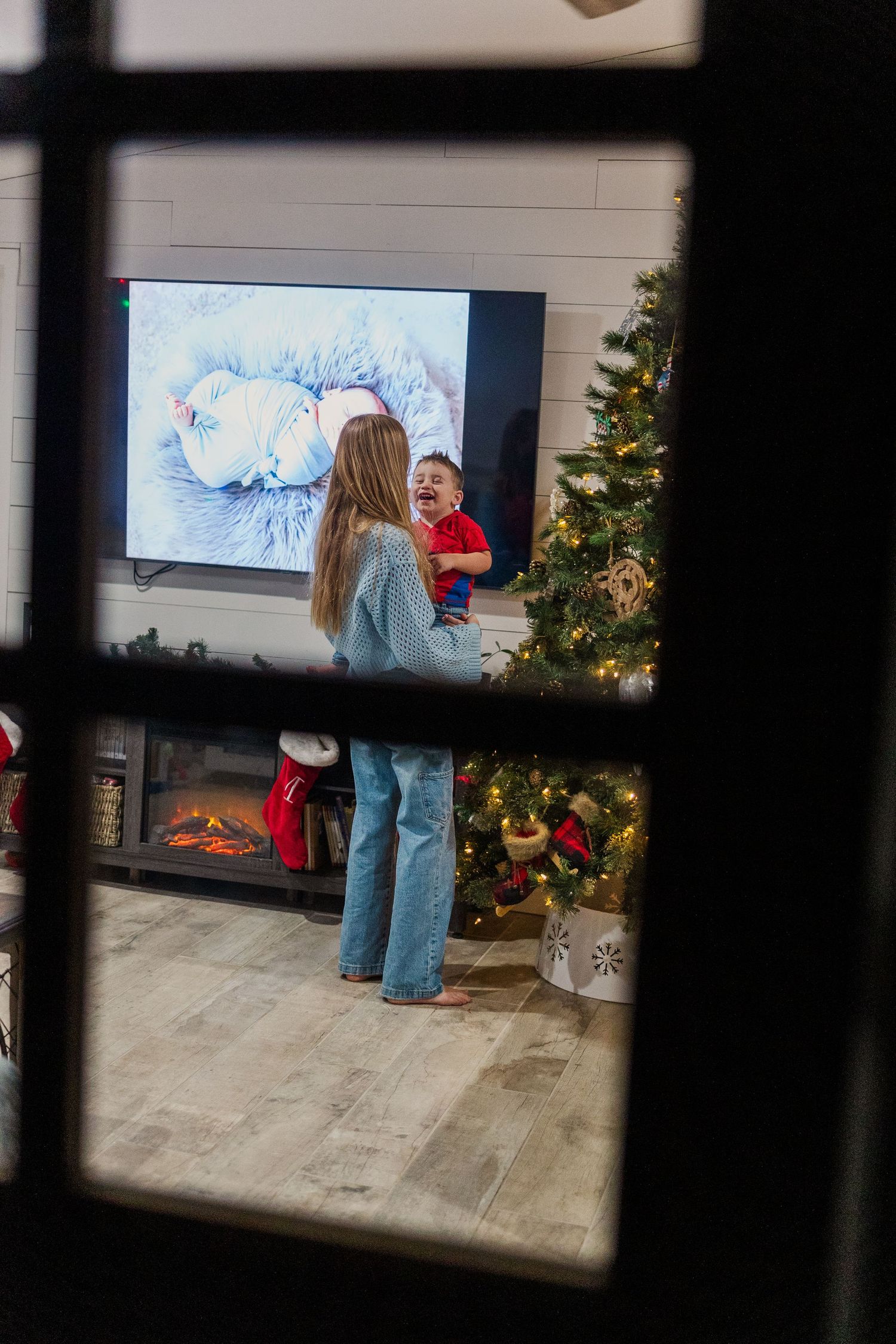 A cozy Christmas scene viewed through a window showing a decorated tree and TV display.