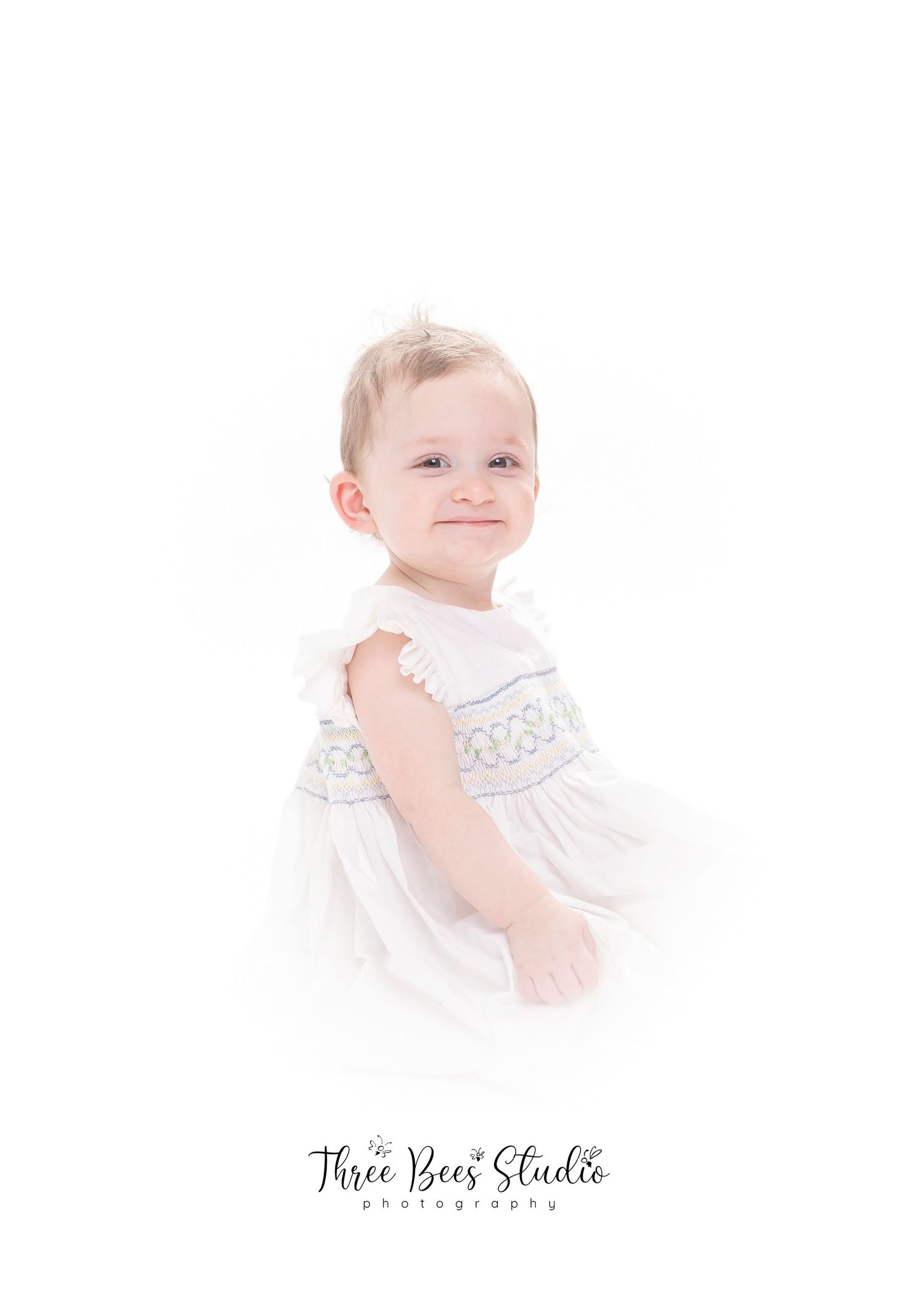 High-key portrait of young child in white smocked dress against light background.