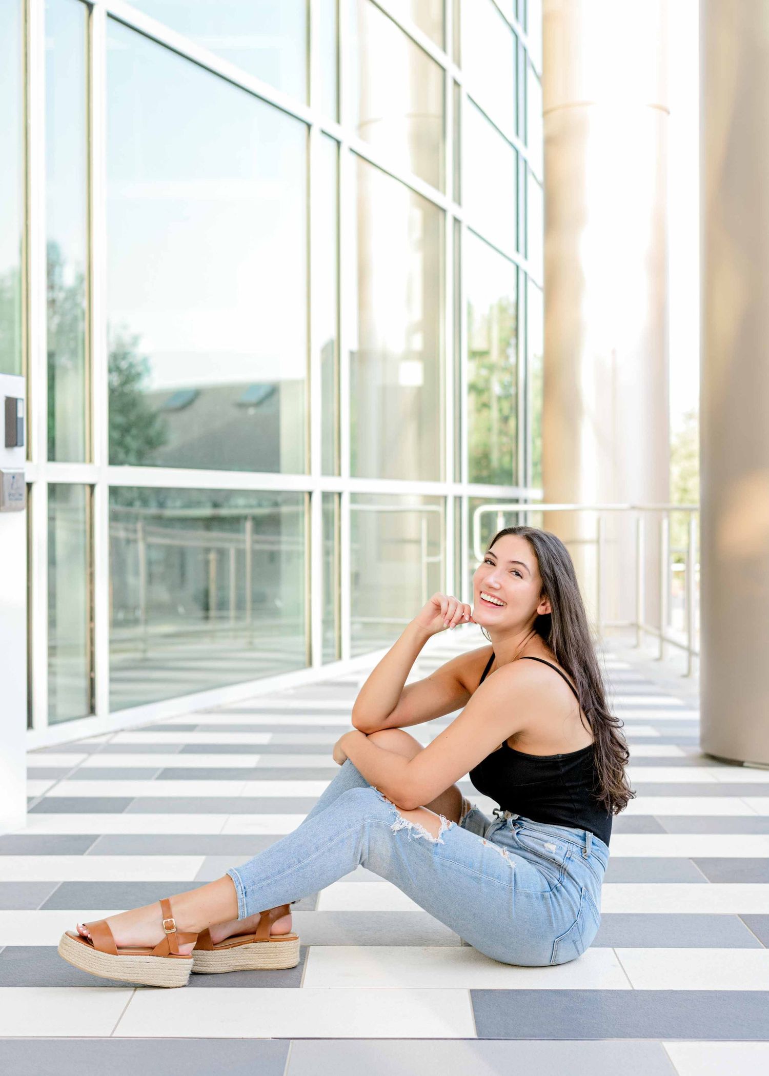 Young woman in casual attire sitting on a striped floor against a glass window wall in an urban setting.