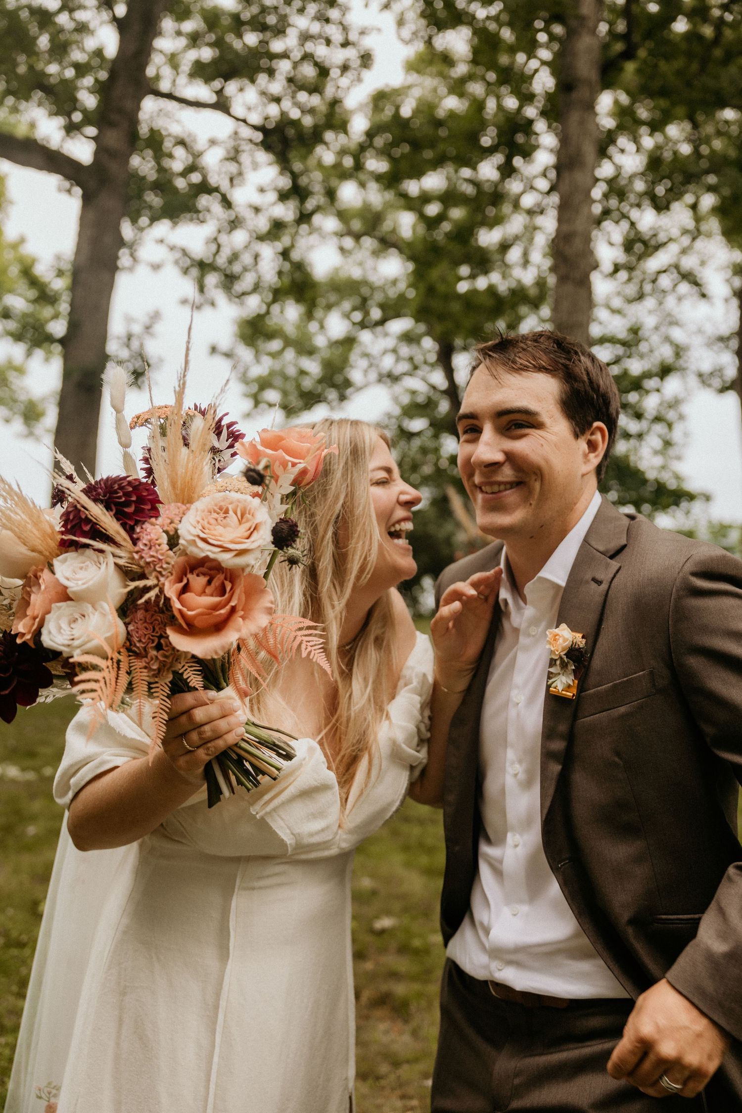 Laughing bride and groom walking outdoors holding colorful boho wedding bouquet among trees.