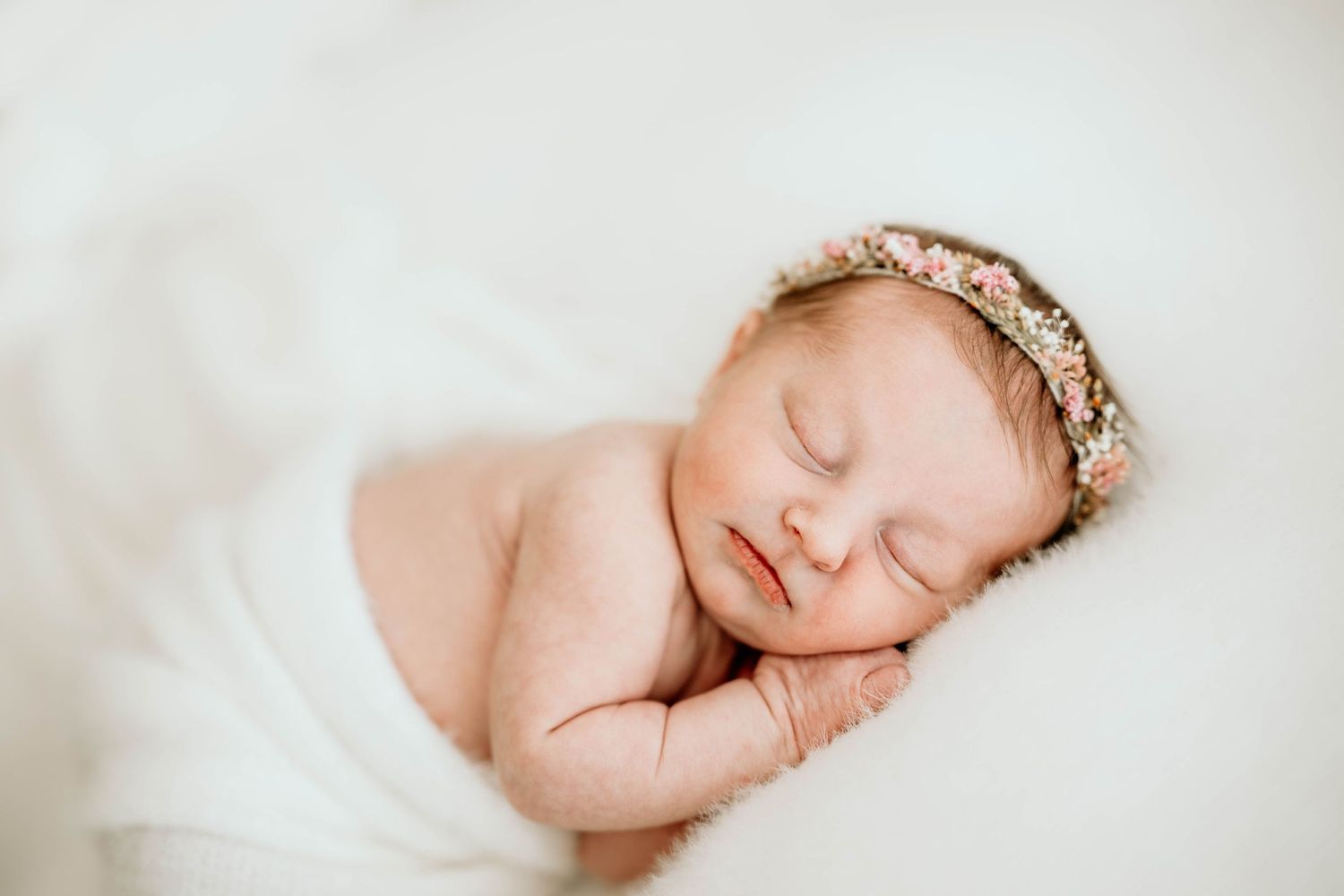 Sleeping newborn wearing delicate lace crown on white blanket.