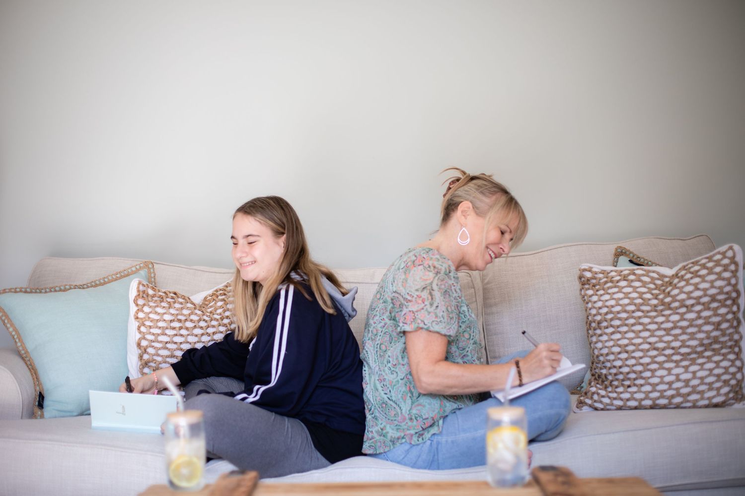 Two people sitting on a gray couch with decorative pillows while writing or studying.
