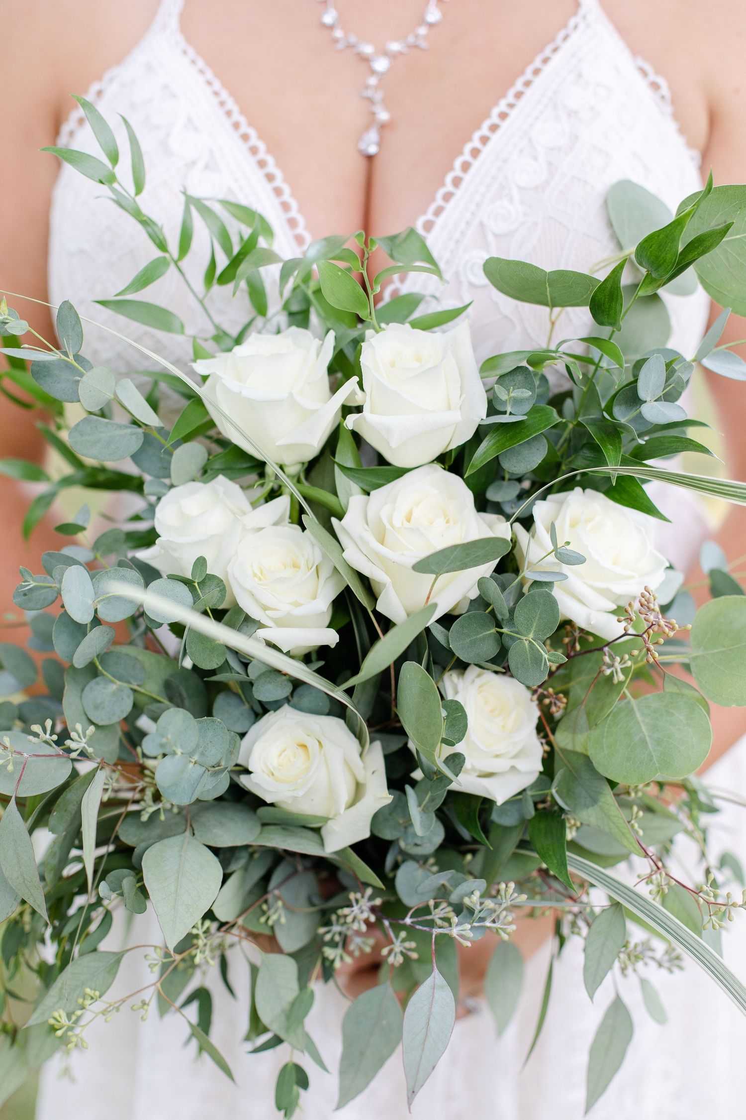 Elegant white rose bridal bouquet with eucalyptus leaves and greenery against a white lace dress background.