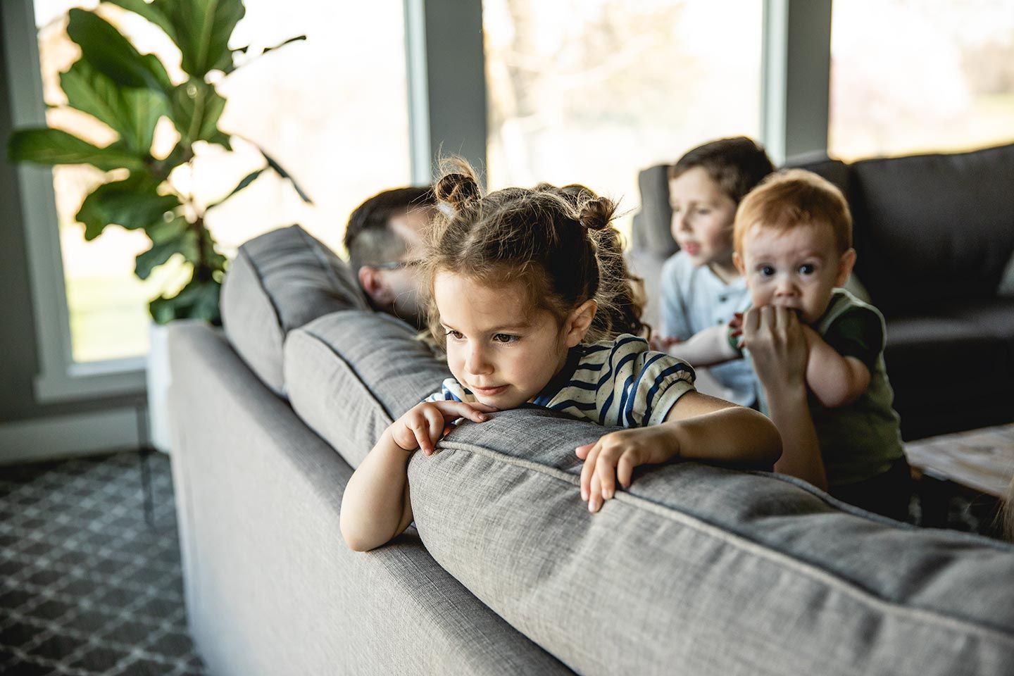 Young girl sitting on the couch with her family.  She is resting on the back of the couch and facing the window.