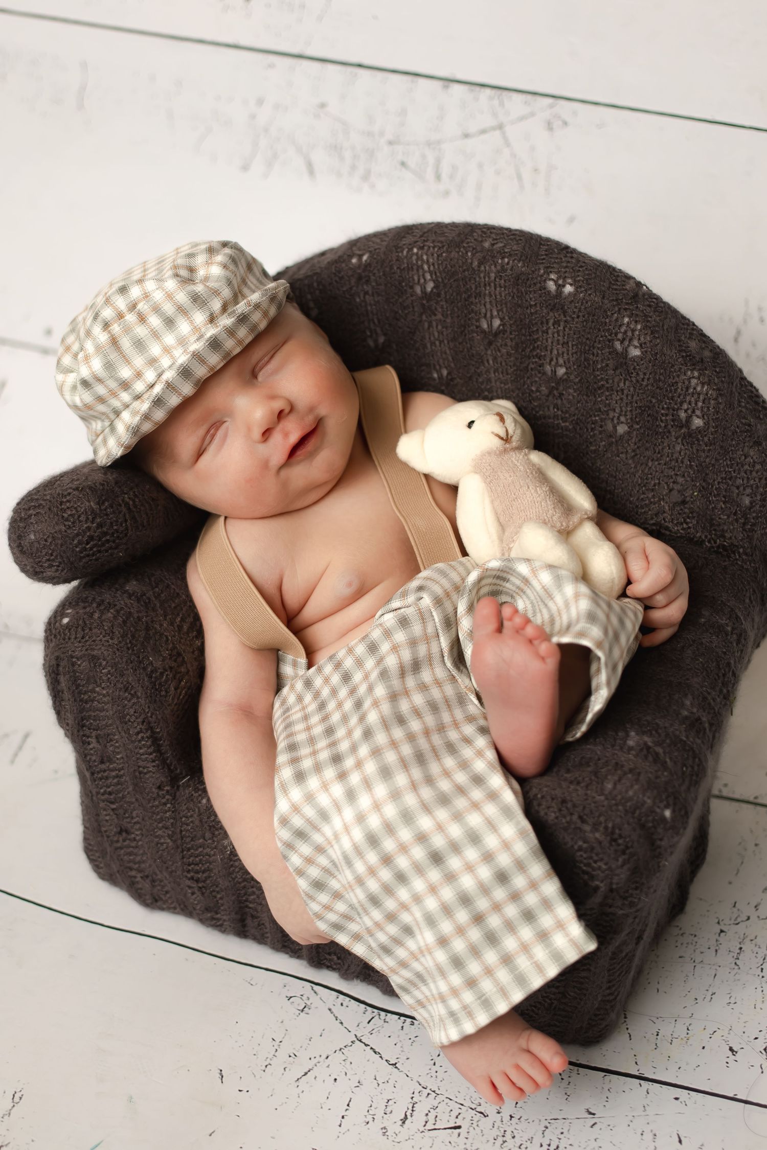 Baby sleeping in gray chair wearing plaid newsboy cap with small stuffed animal.