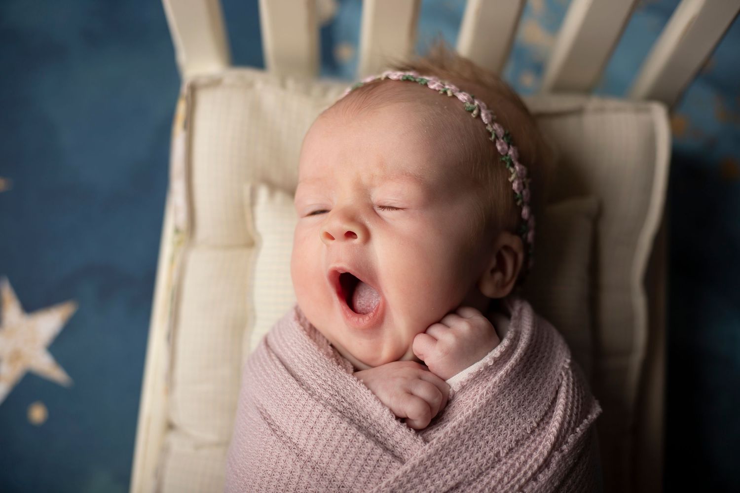 Sleepy newborn yawning while wrapped in pink swaddle on chair with star background.