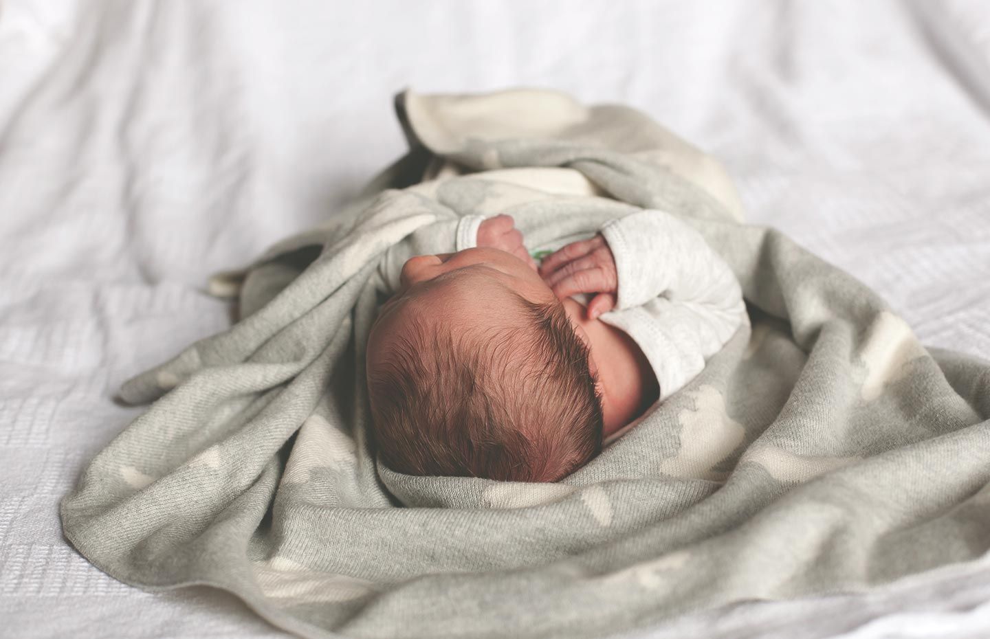 Newborn baby wrapped loosely in blanket and wearing a white onesie, laying on bed