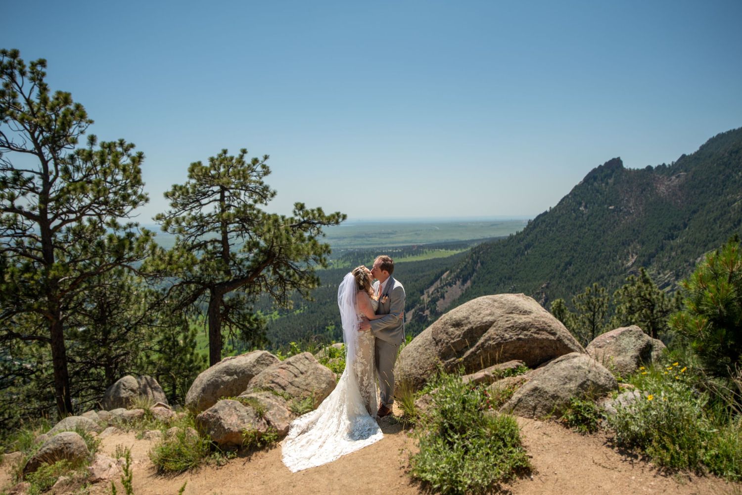 Wedding couple embraces on a rocky mountain overlook with scenic valley vista below.