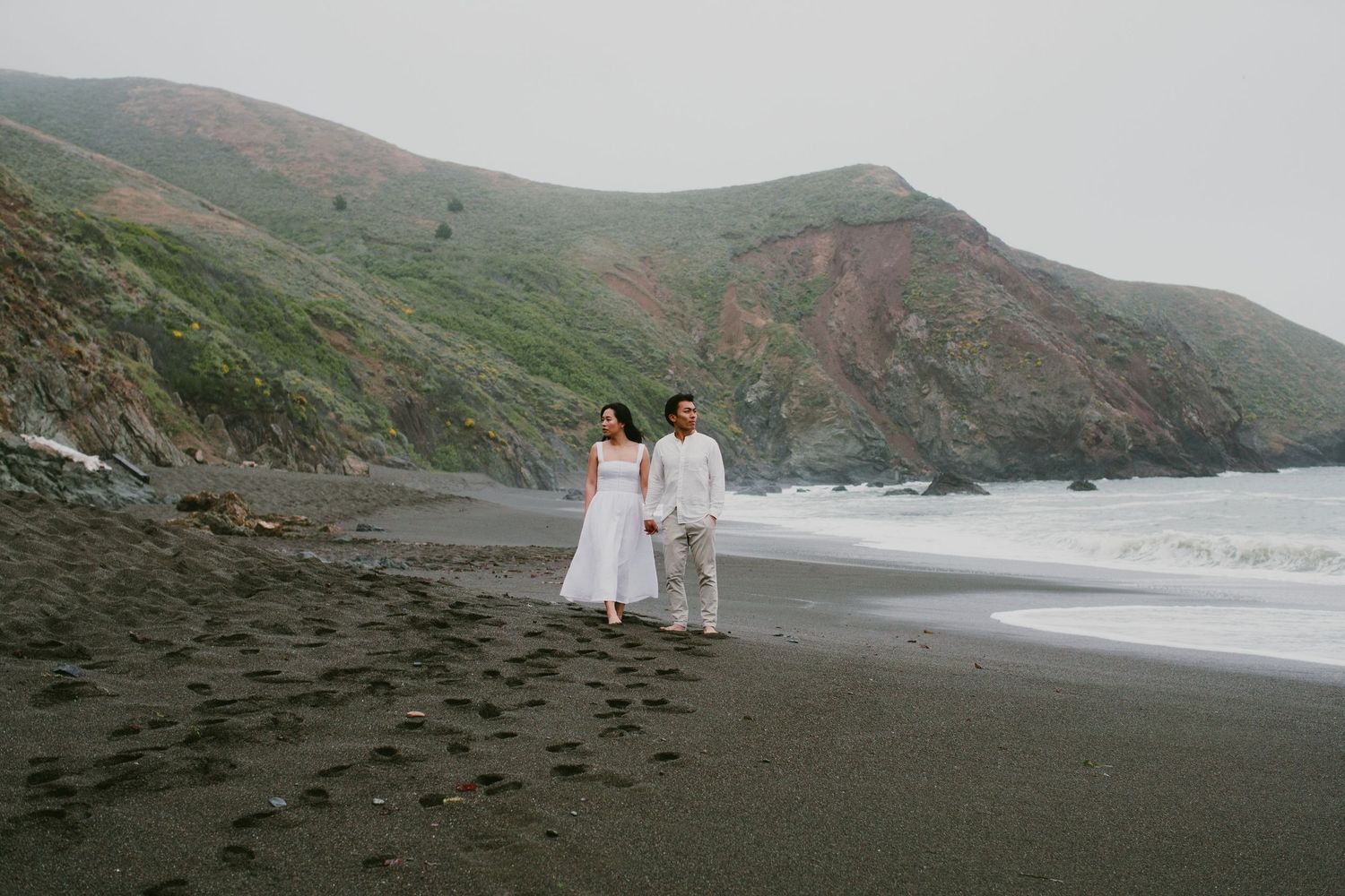 A couple in white outfits walks along a misty black sand beach with cliffs in the background.