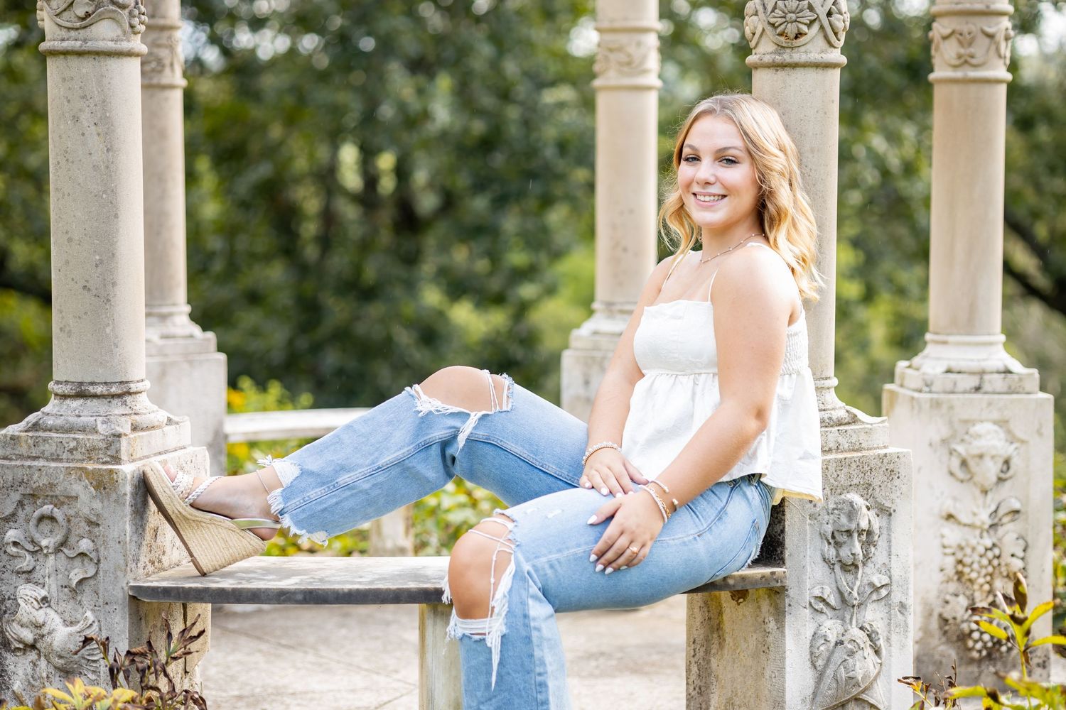 Woman in white top and jeans sitting on stone balustrade in garden setting.