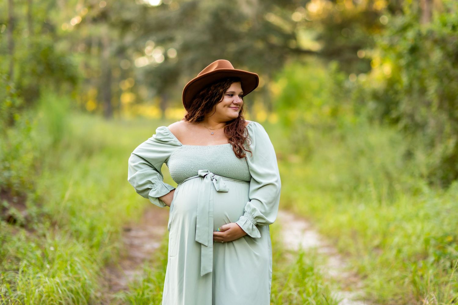 Expecting mother in a mint dress and brown hat poses in a scenic wooded setting.