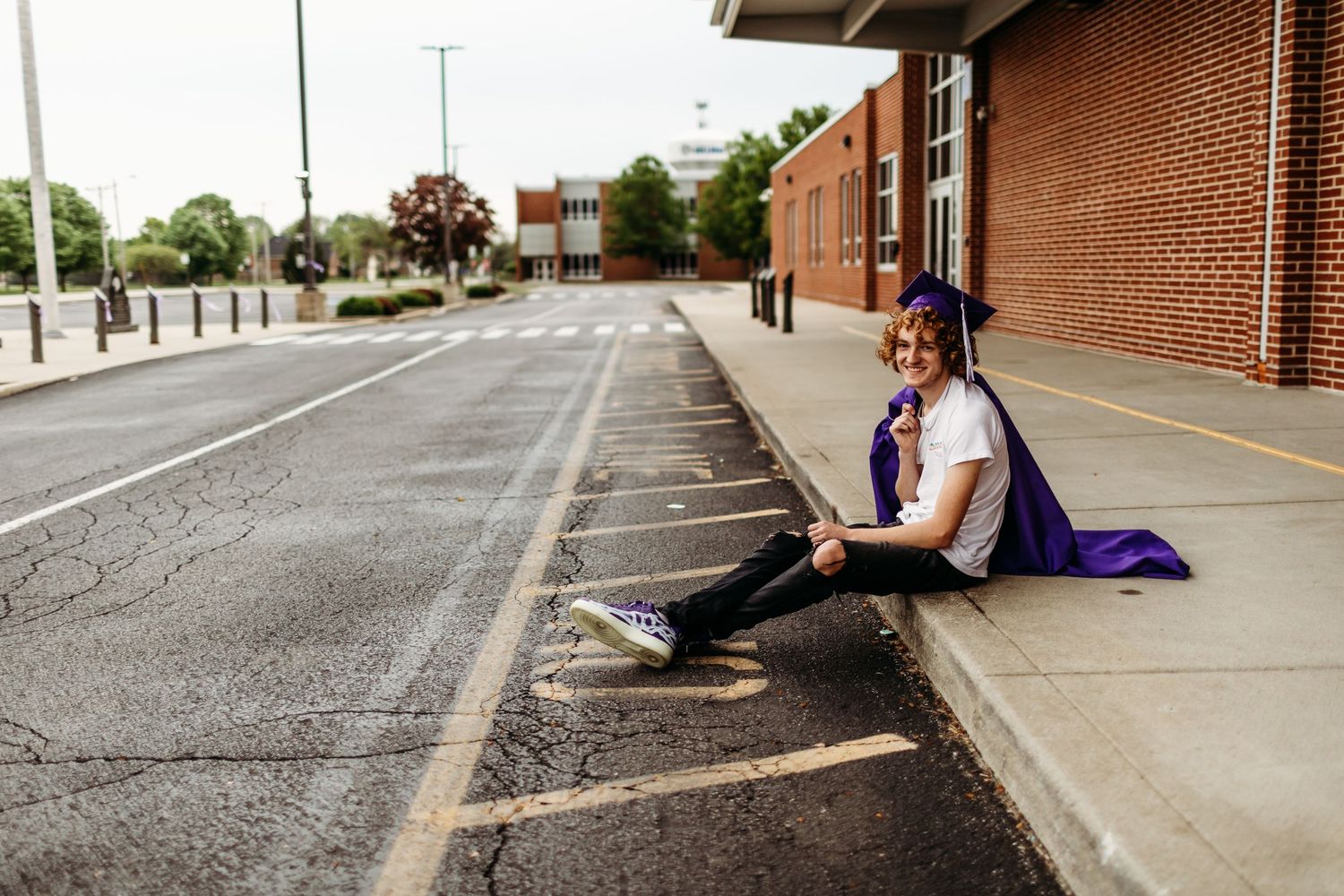 A person sits casually on a sidewalk curb outside a brick building.
