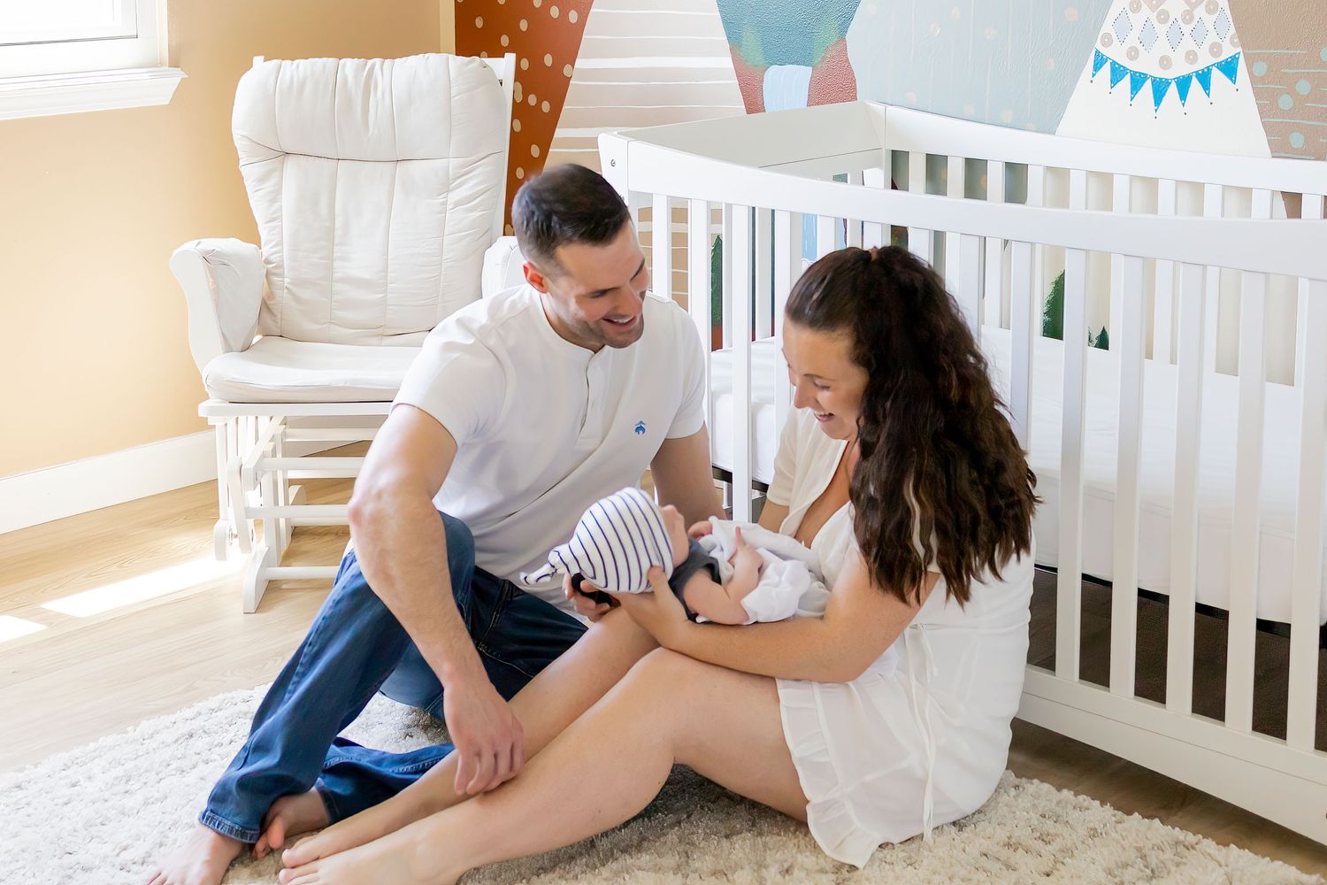 mom and dad sat on floor of nursery, mom holding newborn, dad looking at mom smiling.  Aurora newborn photographer