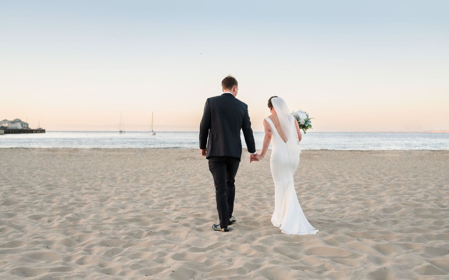 Newlyweds walking along a beach at sunset holding hands.