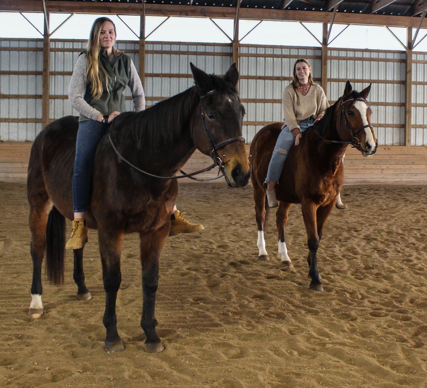 Two horses and riders standing side by side in an indoor riding arena.
