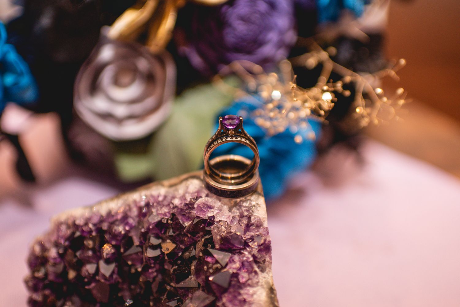 Close-up of wedding rings sitting atop dark purple crystal with blurred floral background.