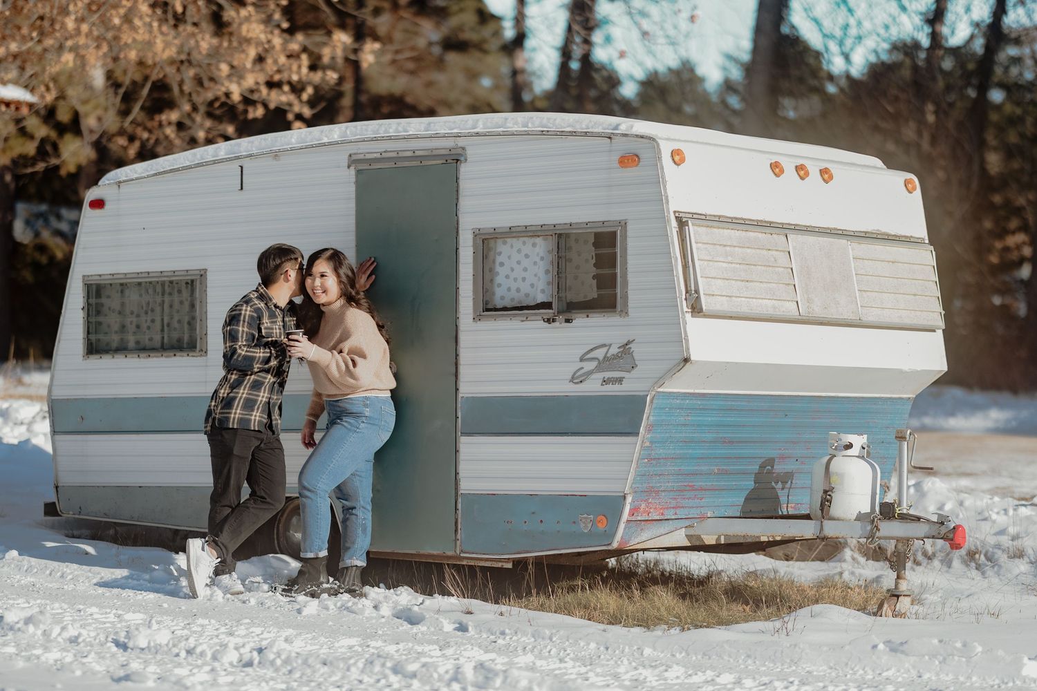 Couple poses casually in front of a vintage blue and white camper trailer in snowy weather.