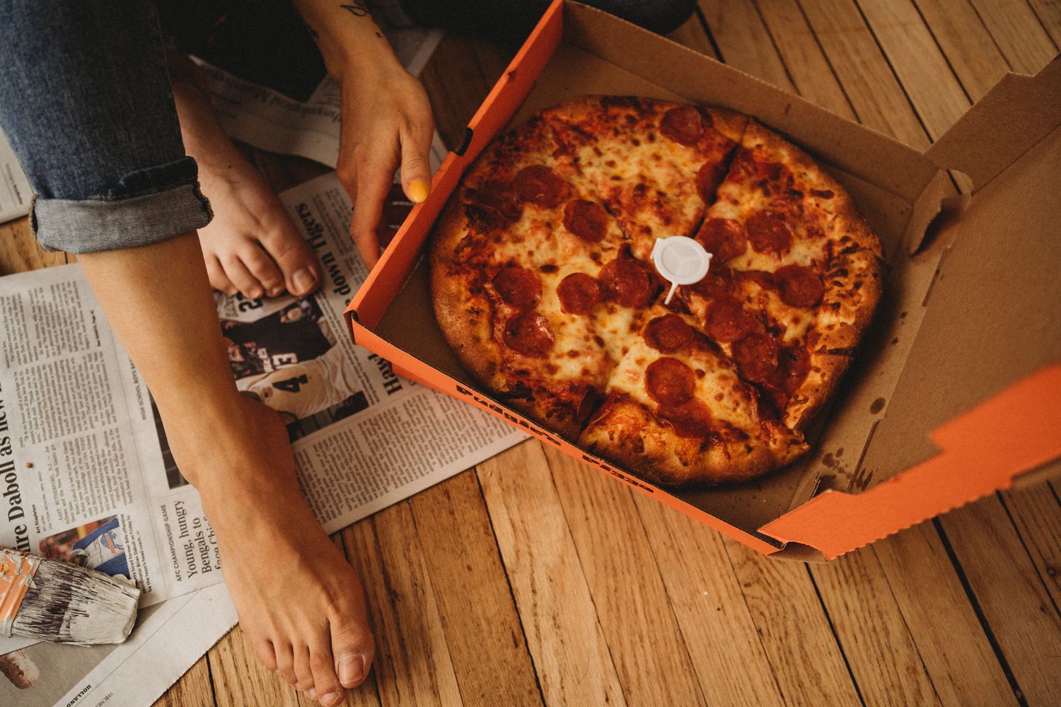 Overhead view of a fresh pizza in delivery box with newspaper and drink on wooden floor.