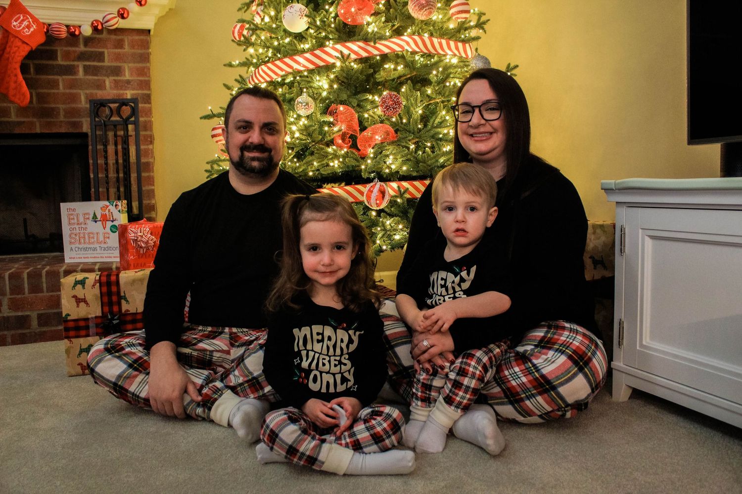 A family wearing matching plaid pajamas poses in front of a decorated Christmas tree with presents.