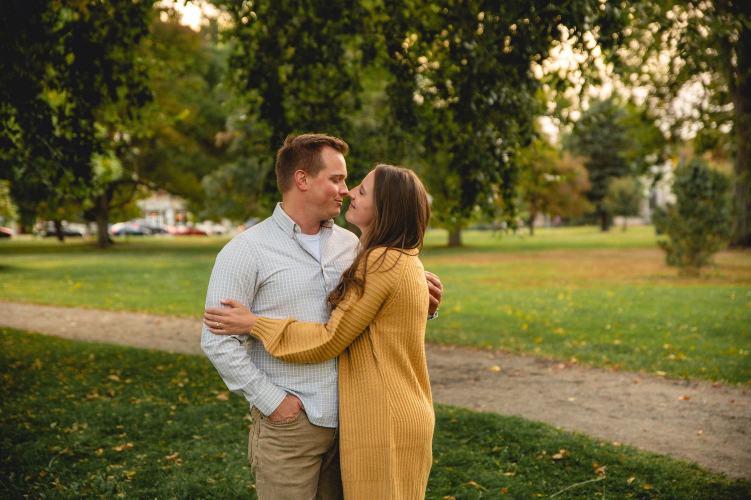 A couple shares an intimate moment in a park with autumn trees in the background.