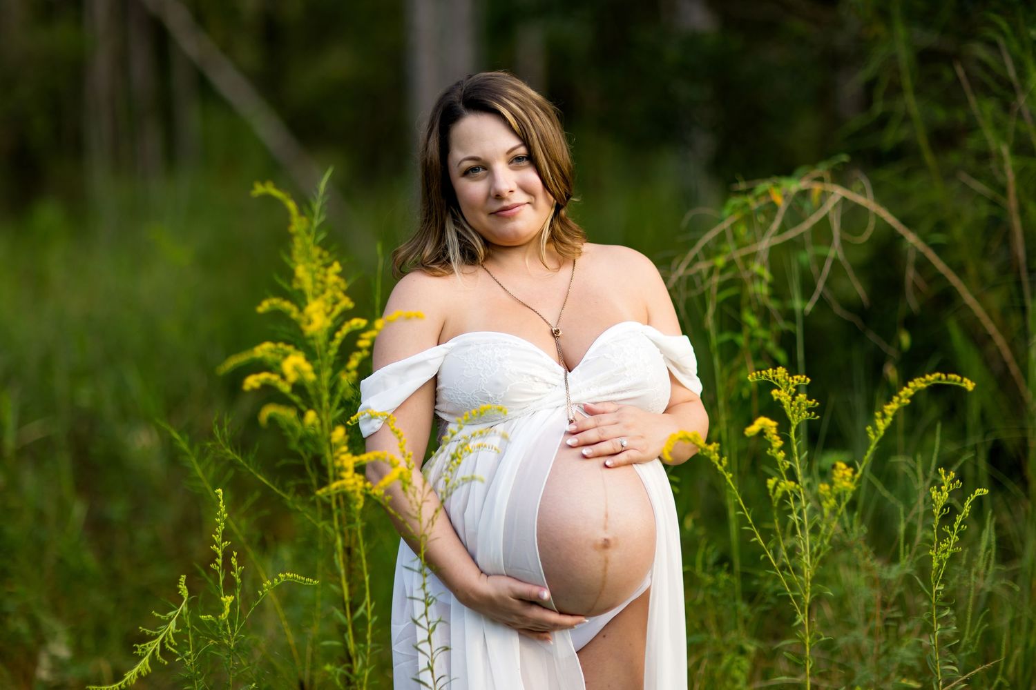 Maternity photos in white dress against lush green foliage background.