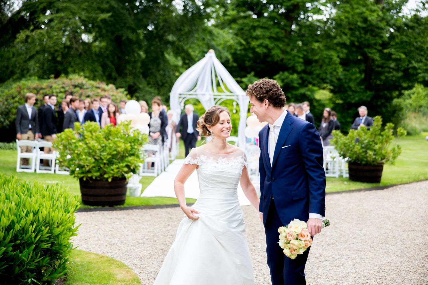 A wedding ceremony setup outdoors with a white arch, greenery, and guests seated on white chairs on a garden path.