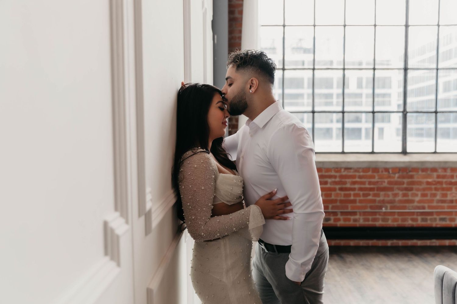 Intimate couple moment captured against white interior wall and window.