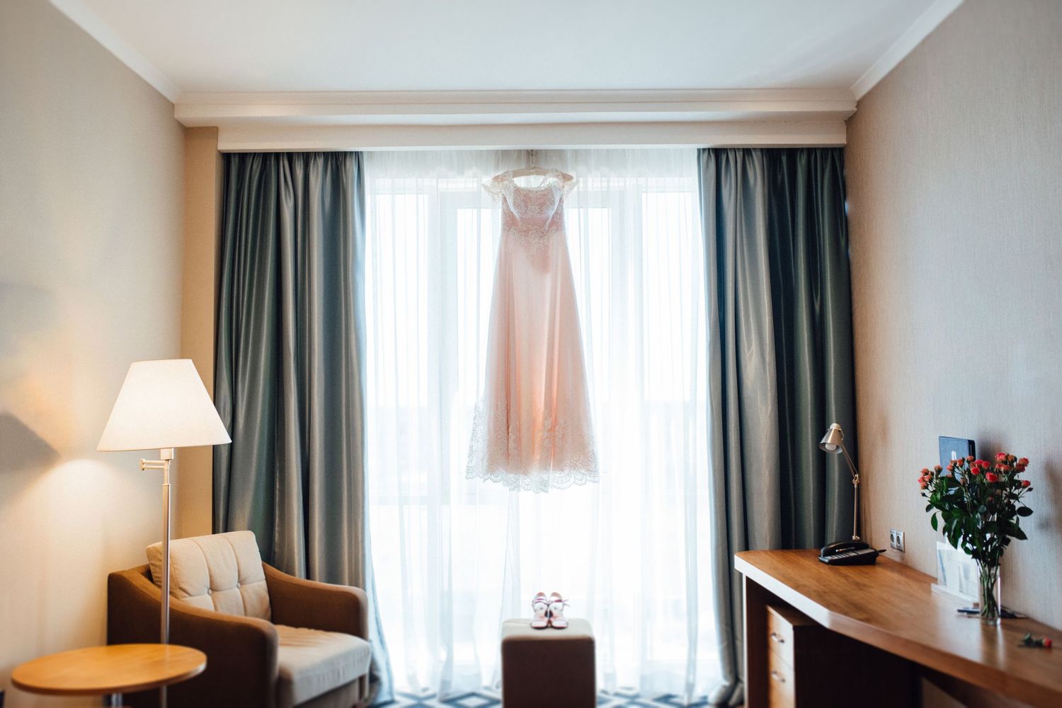 Blush wedding dress hanging in hotel room window with teal curtains and warm lighting.