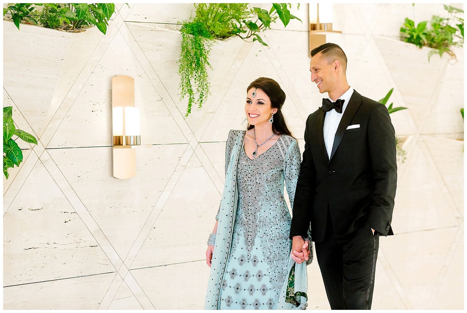 Formal couple in traditional and cultural wedding attire pose against white geometric wall with hanging plants.