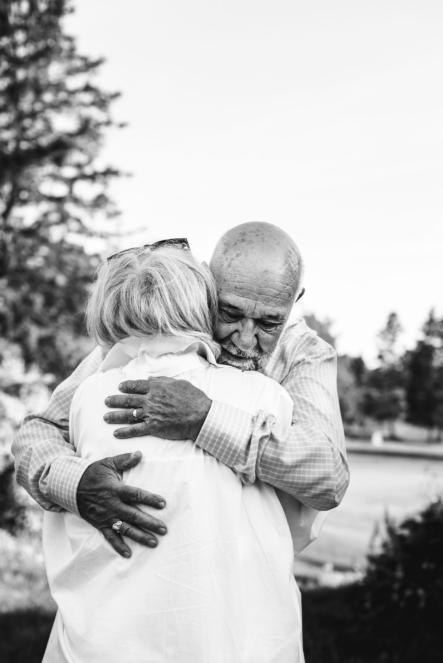 Black and white image of a caucasian man facing the camera as he hugs a woman in a white shirt with white hair.
