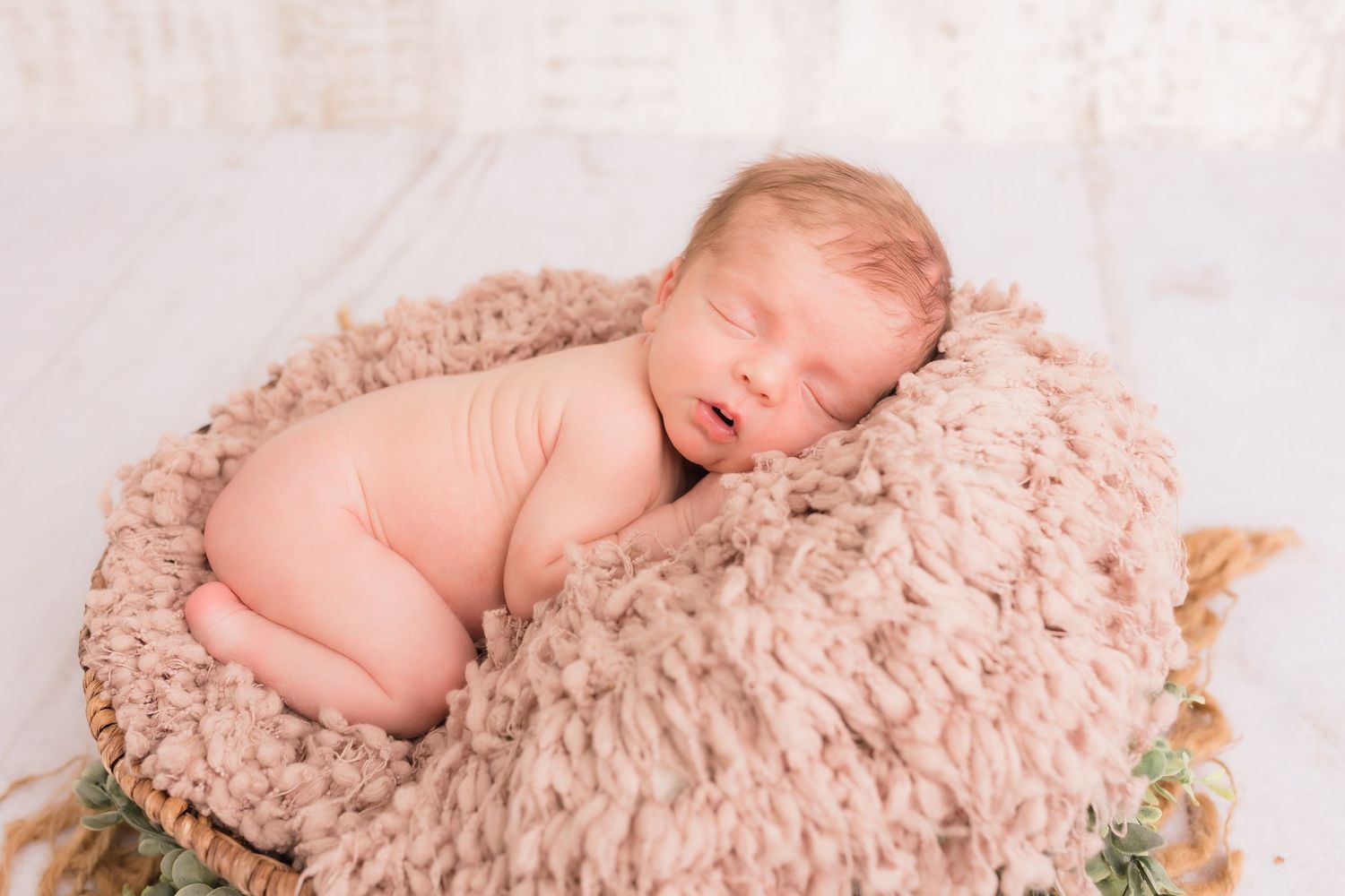 Sleeping newborn baby curled up in a round wicker basket with soft brown fuzzy filler during a studio photo session.
