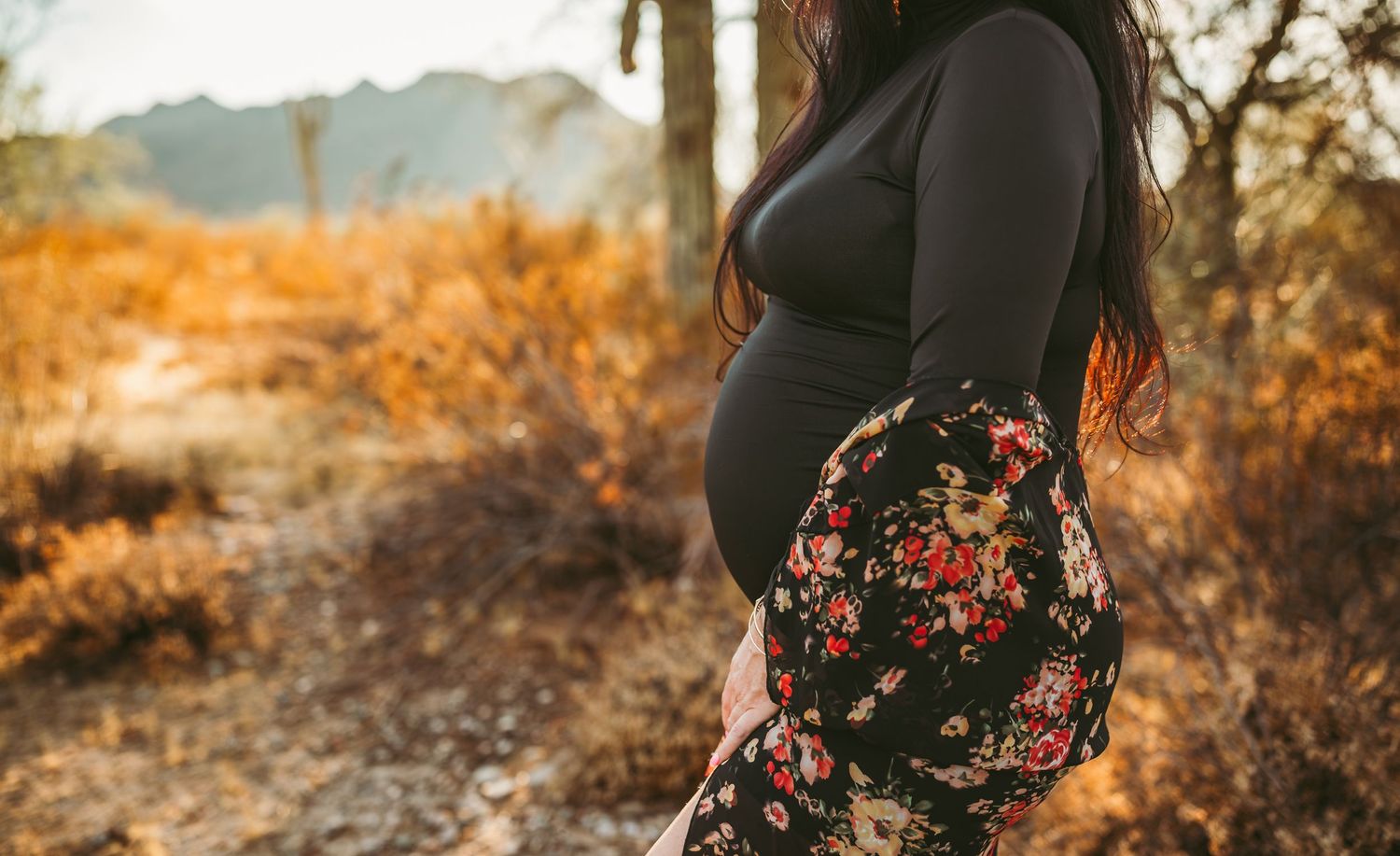 a pregnant women stands holding her belly wearing a black bodysuit with floral accents
