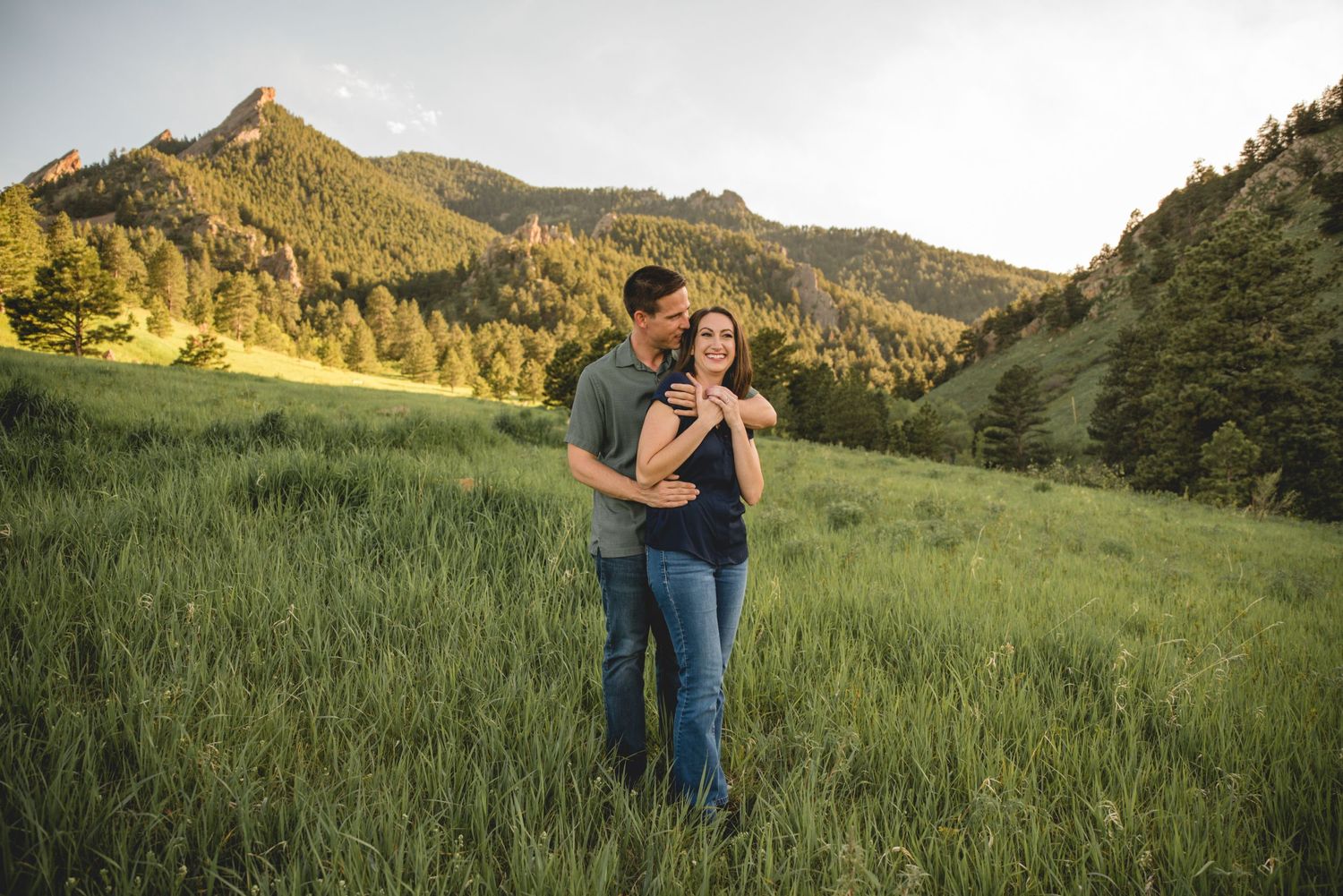 A couple shares an intimate moment in a grassy meadow with mountain peaks in the backdrop.