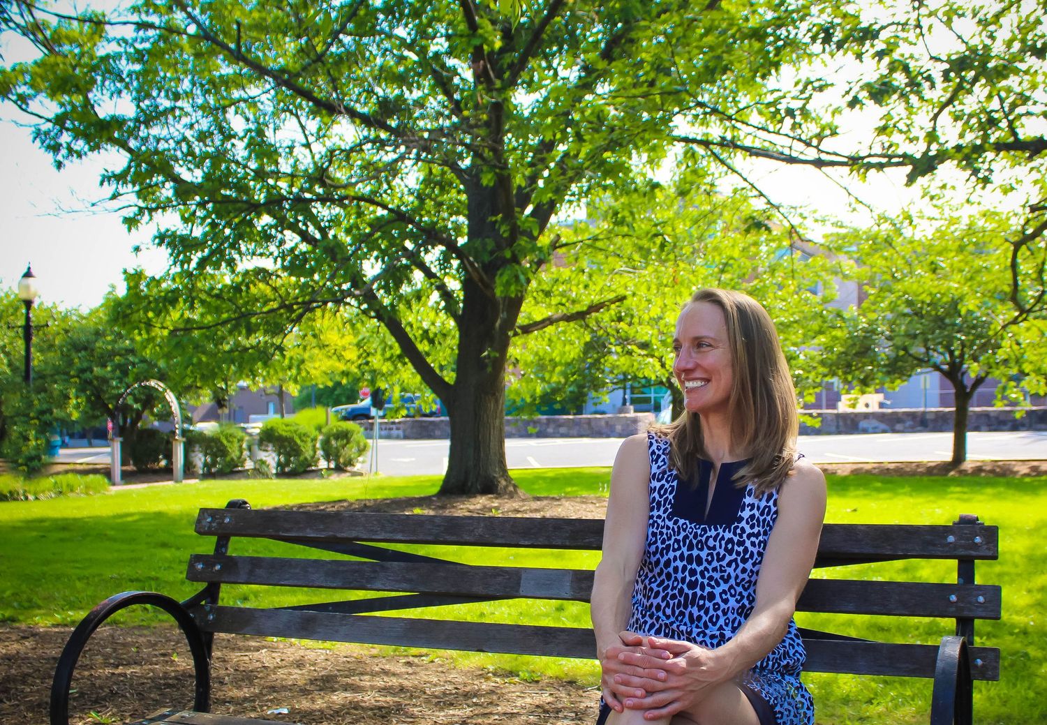 A person in a blue patterned dress sits on a wooden bench under a large tree in a park.