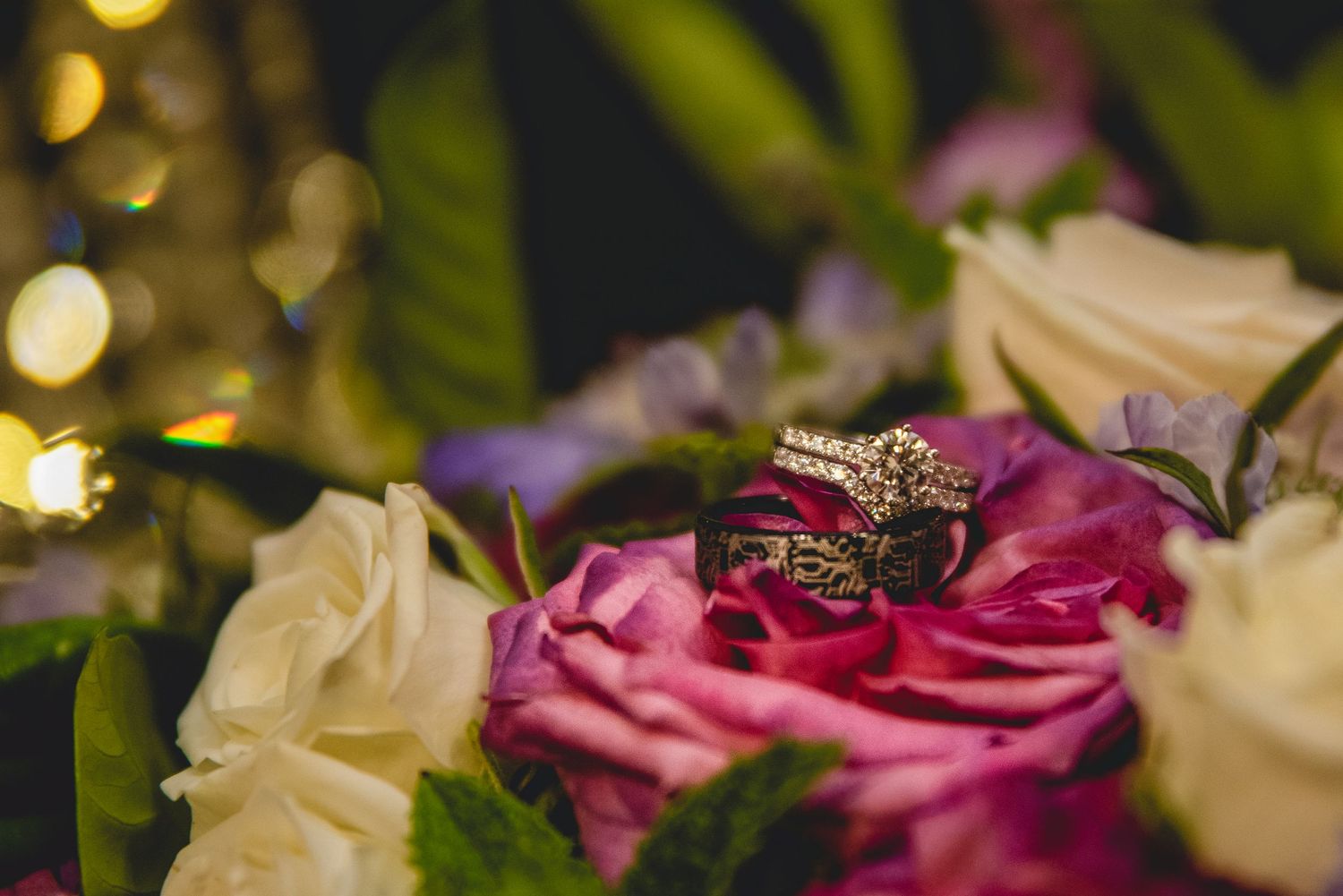 Diamond engagement rings displayed on pink and white roses with bokeh lights in background.