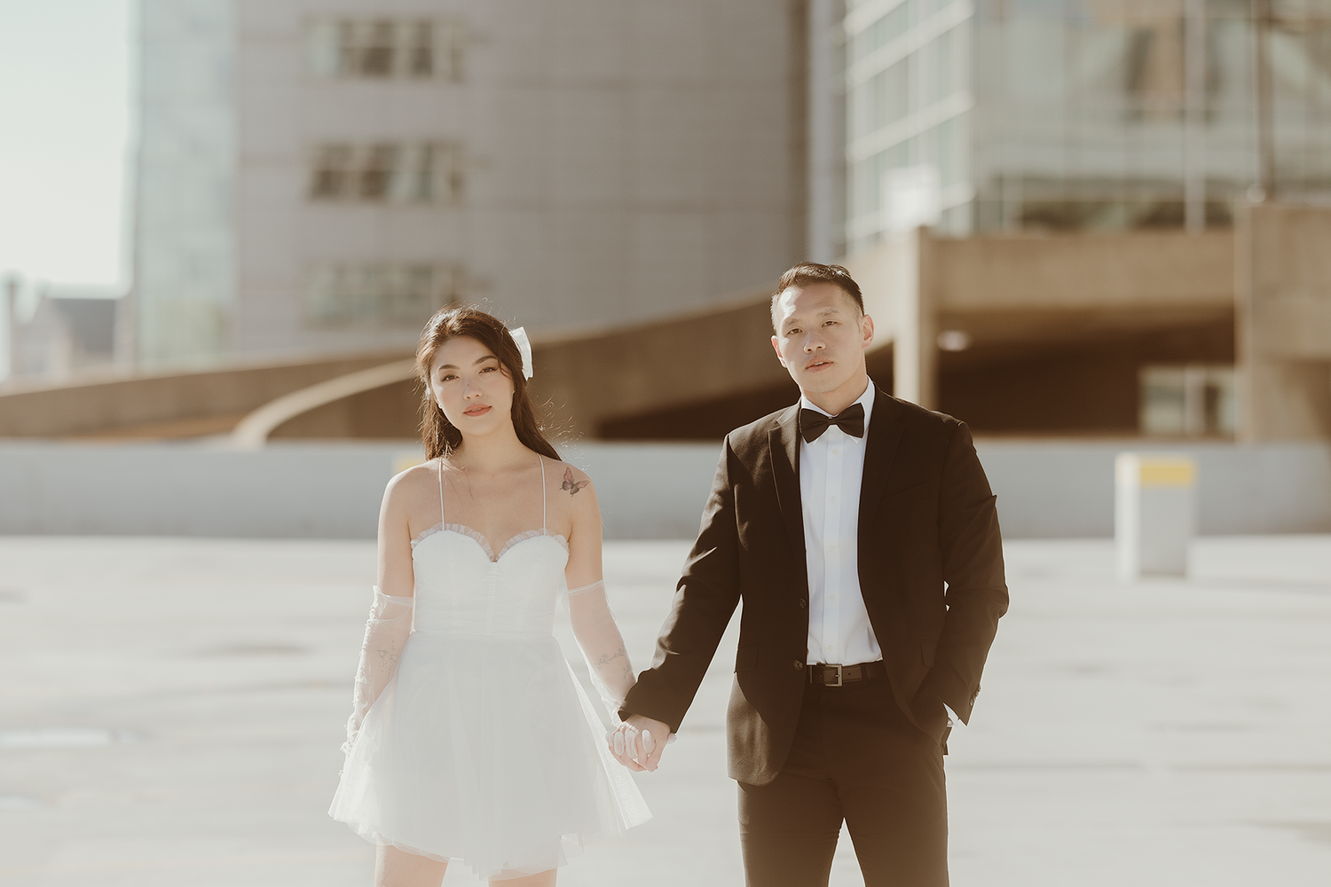 Wedding couple in formal attire poses against modern architecture during sunset photoshoot.