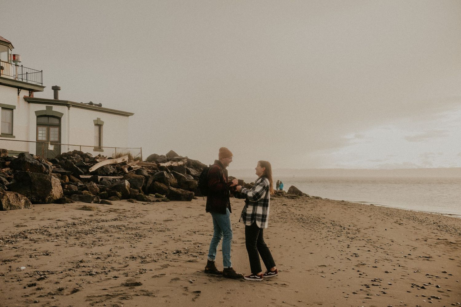 A couple shares a tender moment while standing together on a cloudy beach near a lighthouse.