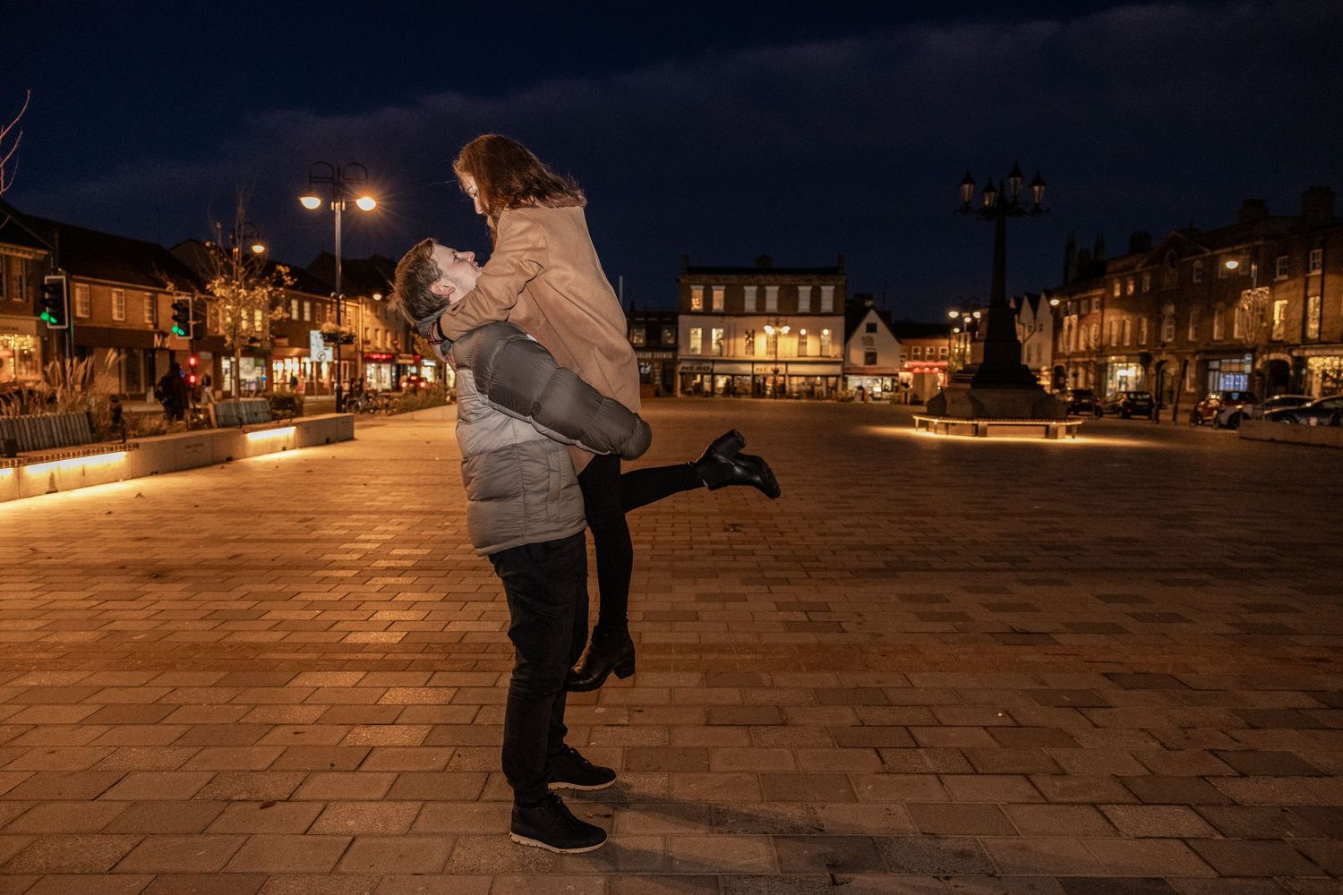 A couple dances together in a romantic evening moment on an illuminated city square.