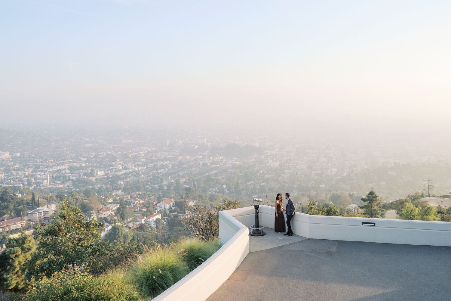 Scenic cityscape view from a hilltop overlook with people silhouetted against a hazy sky.