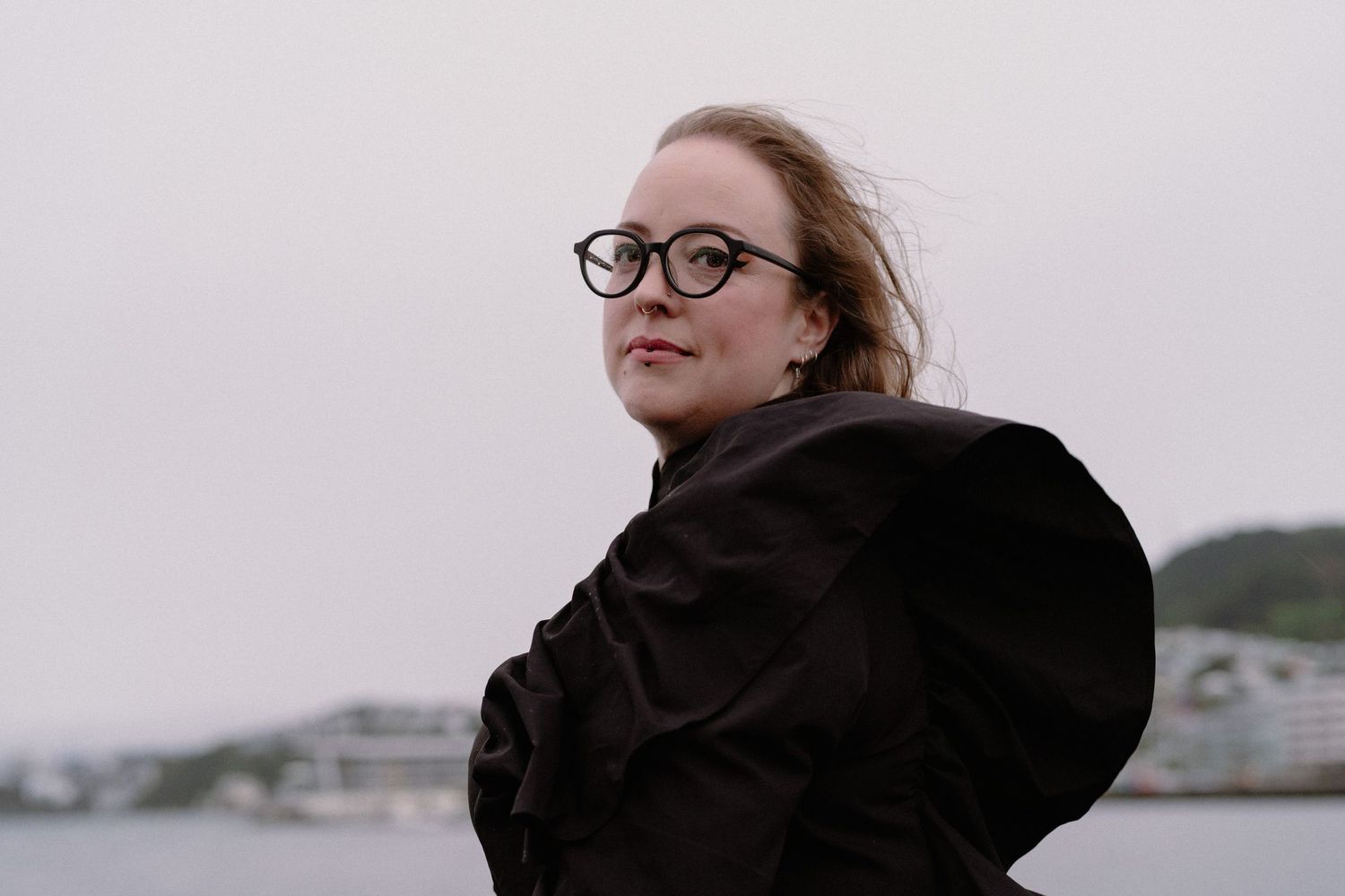 Emma Barnes stands on the Wellington waterfront wearing a black dress and black rim glasses. The wind is blowing her hair and dress