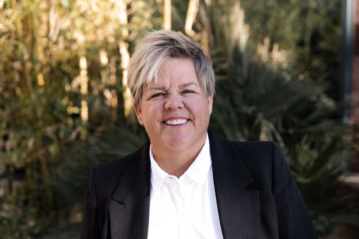 A person in a black blazer and white shirt smiles warmly while posing outdoors in natural lighting.