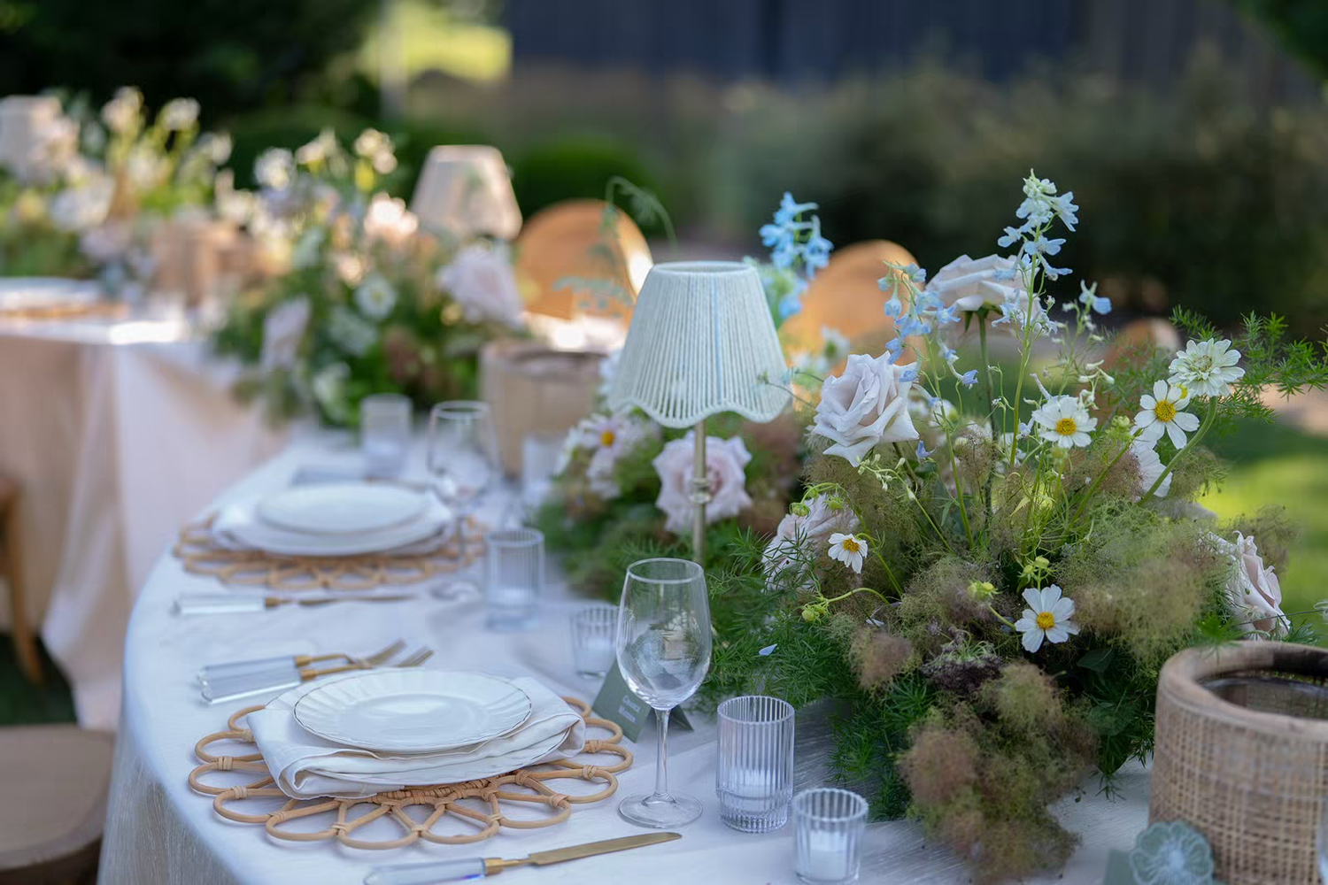 An elegant outdoor table setting with white linens and floral centerpieces extends along a garden path at sunset.