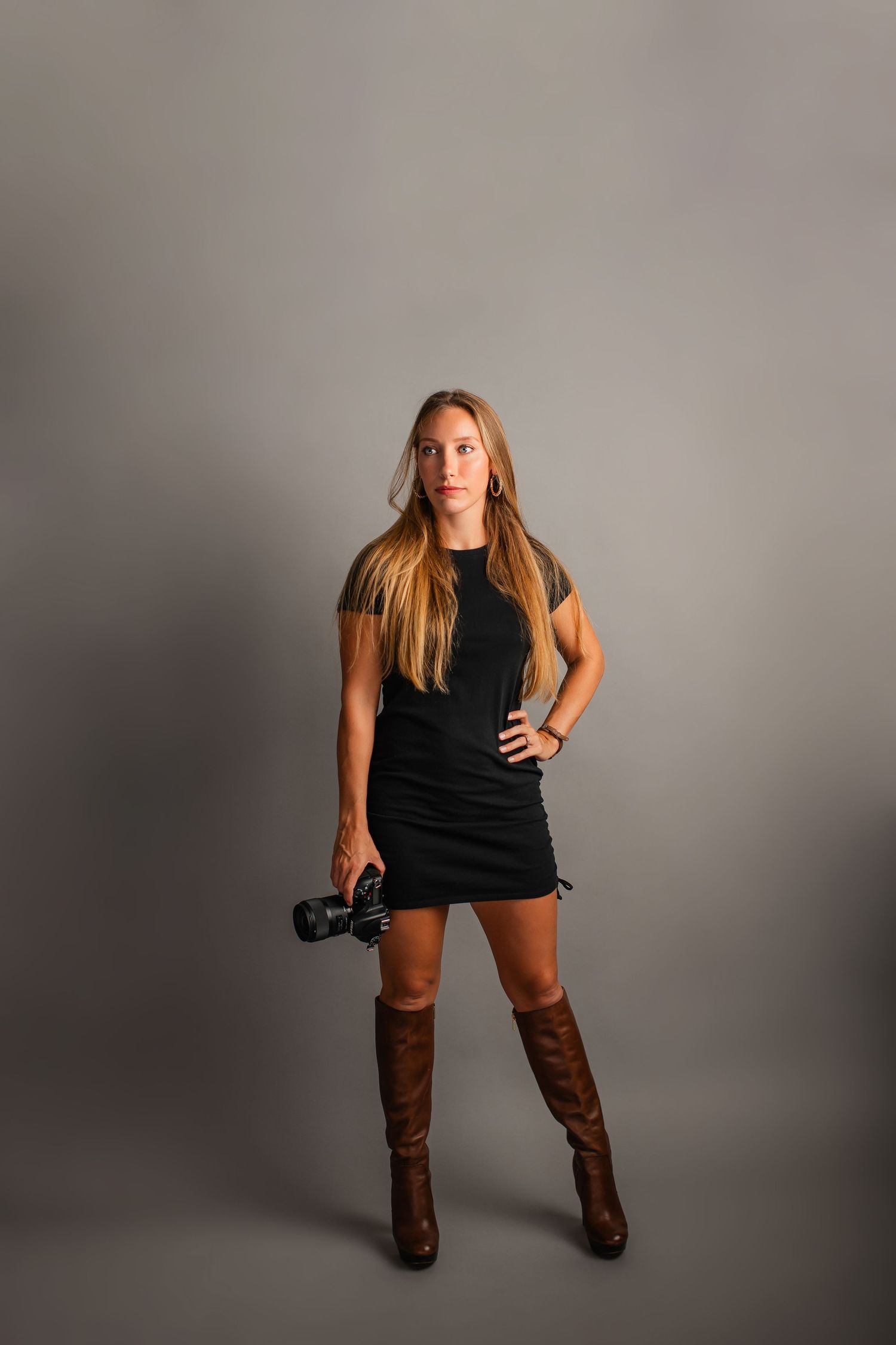 A photographer in a black mini dress and brown knee-high boots poses with a camera against a gray studio backdrop.