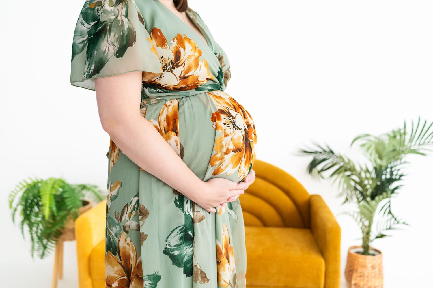 profile of baby bump from shoulder down; mom in green + yellow floral dress; yellow couch and greenery in background. in studio maternity
