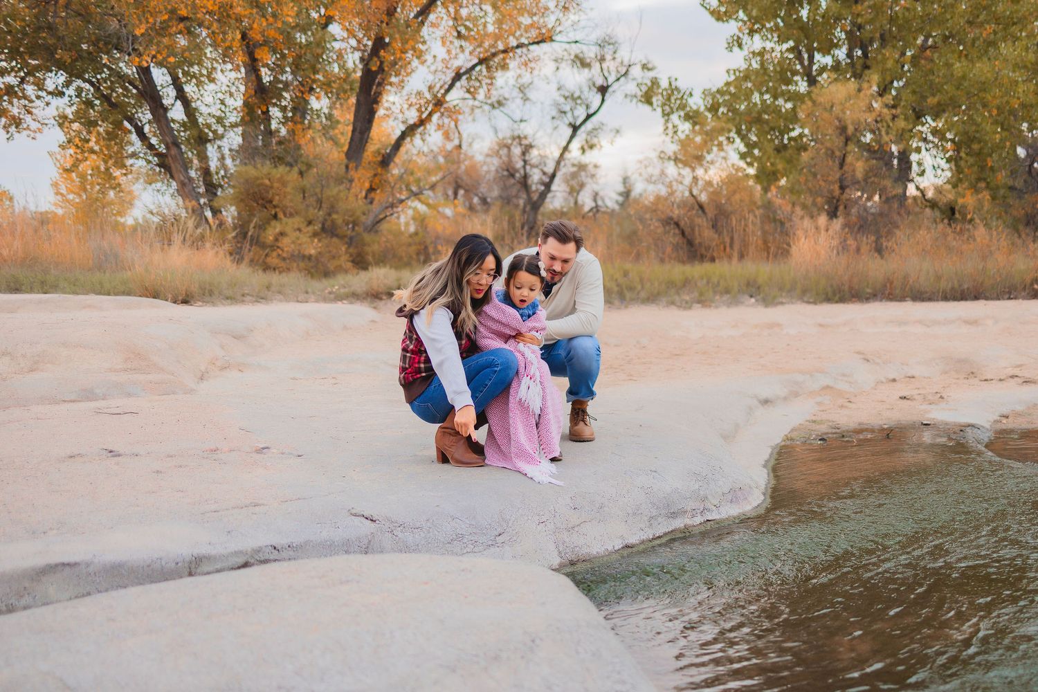 Family of three crouching on rocky riverbank surrounded by autumn trees in golden fall colors.  Mom, Dad and girl aged around 4