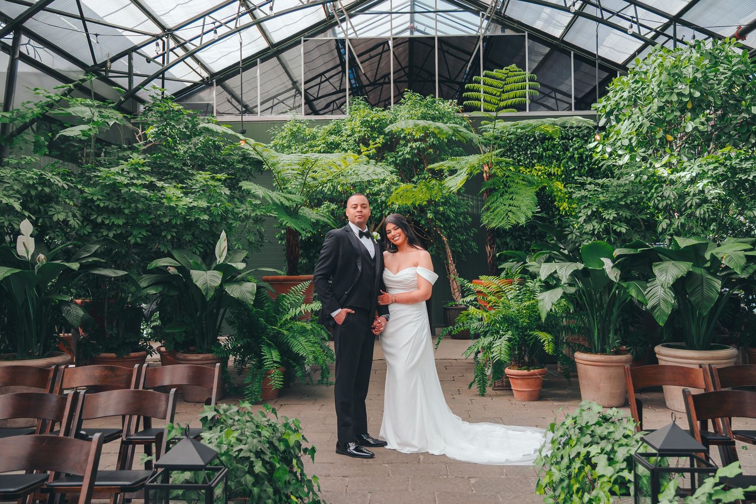 Elegant wedding couple walks along glass-walled building surrounded by lush greenery at dusk.