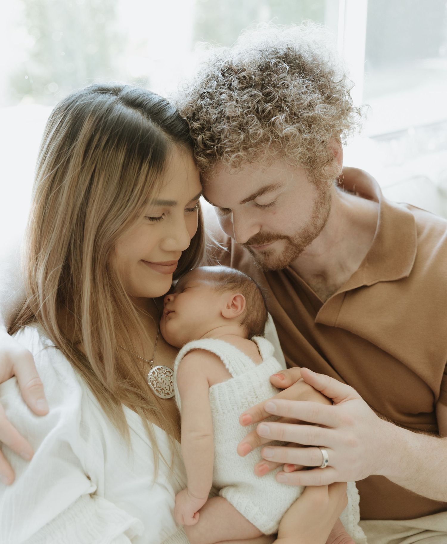 A couple stands by a window in soft light, tenderly holding their newborn baby in a peaceful moment at home.