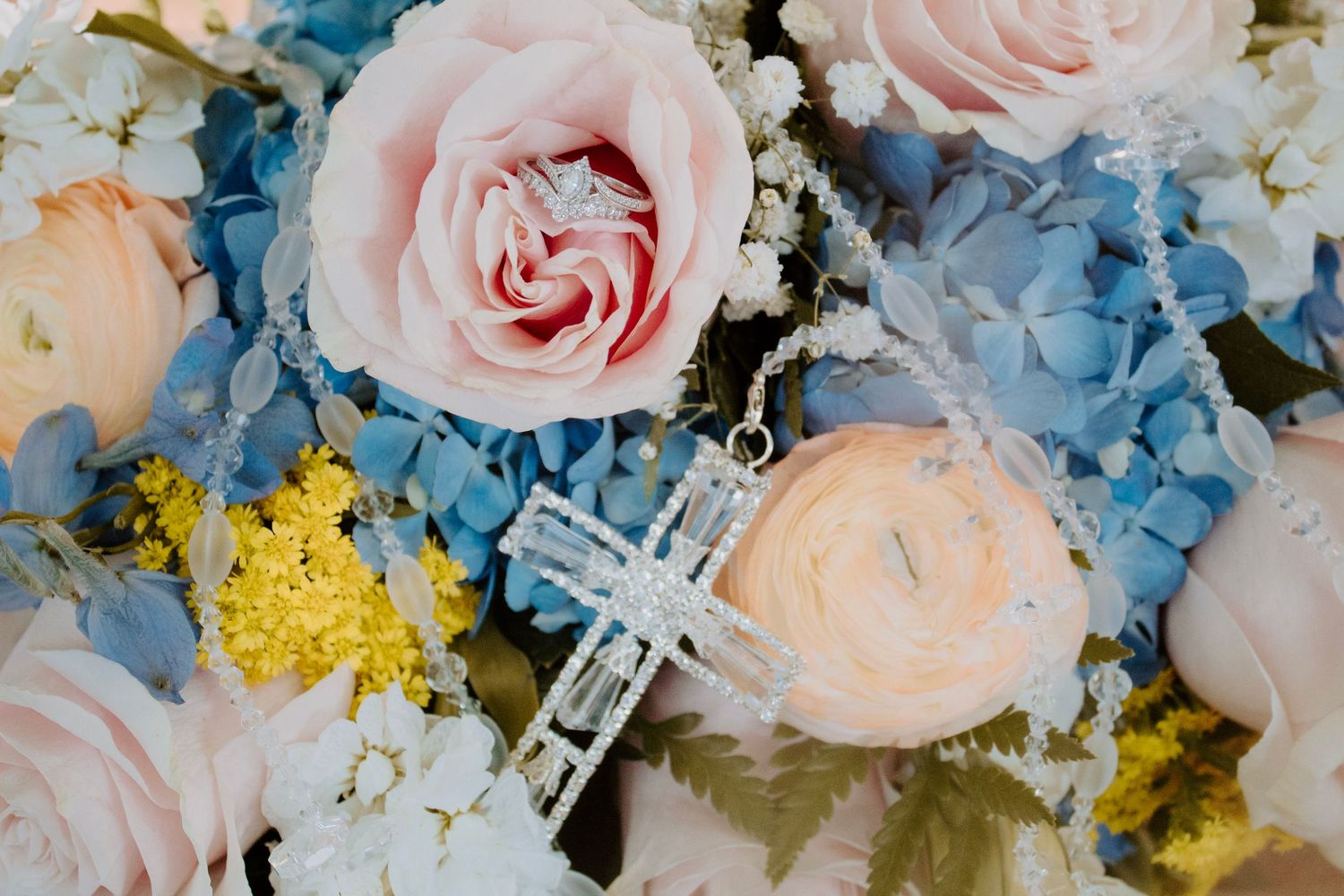 Silver cross necklace resting on pink roses and blue hydrangeas wedding bouquet.