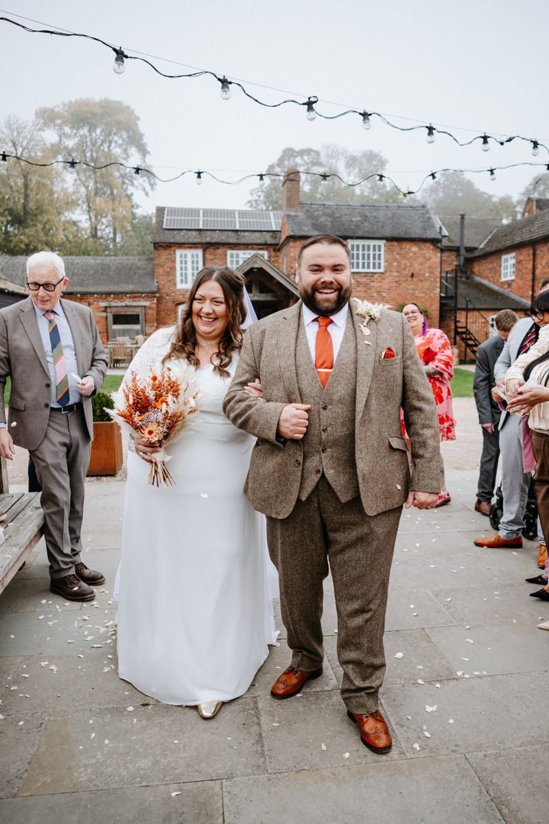 Joyful wedding couple walking through confetti and bubbles as guests celebrate outside a rustic brick venue.