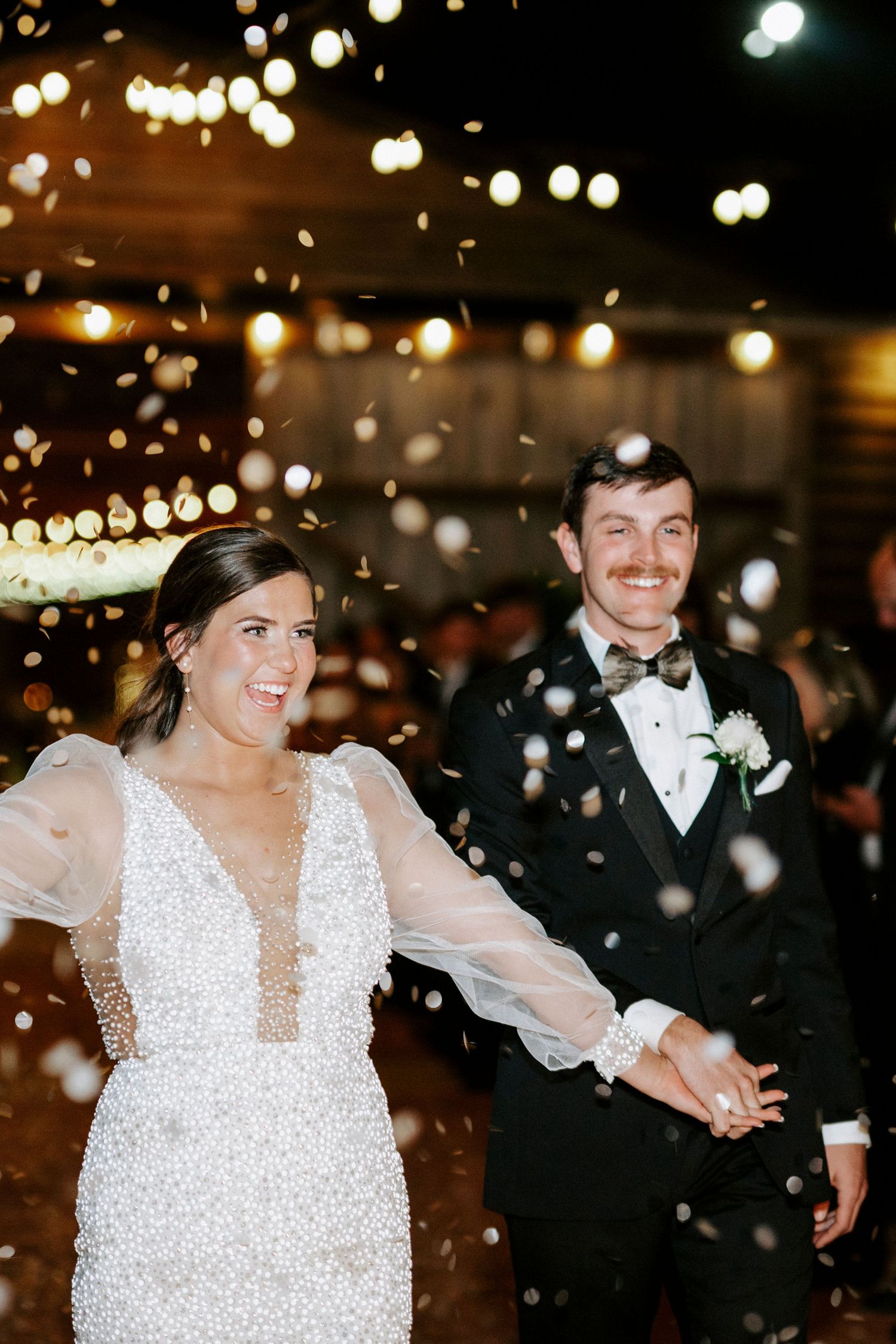 A joyful couple dances together at a wedding reception under twinkling lights and falling confetti.